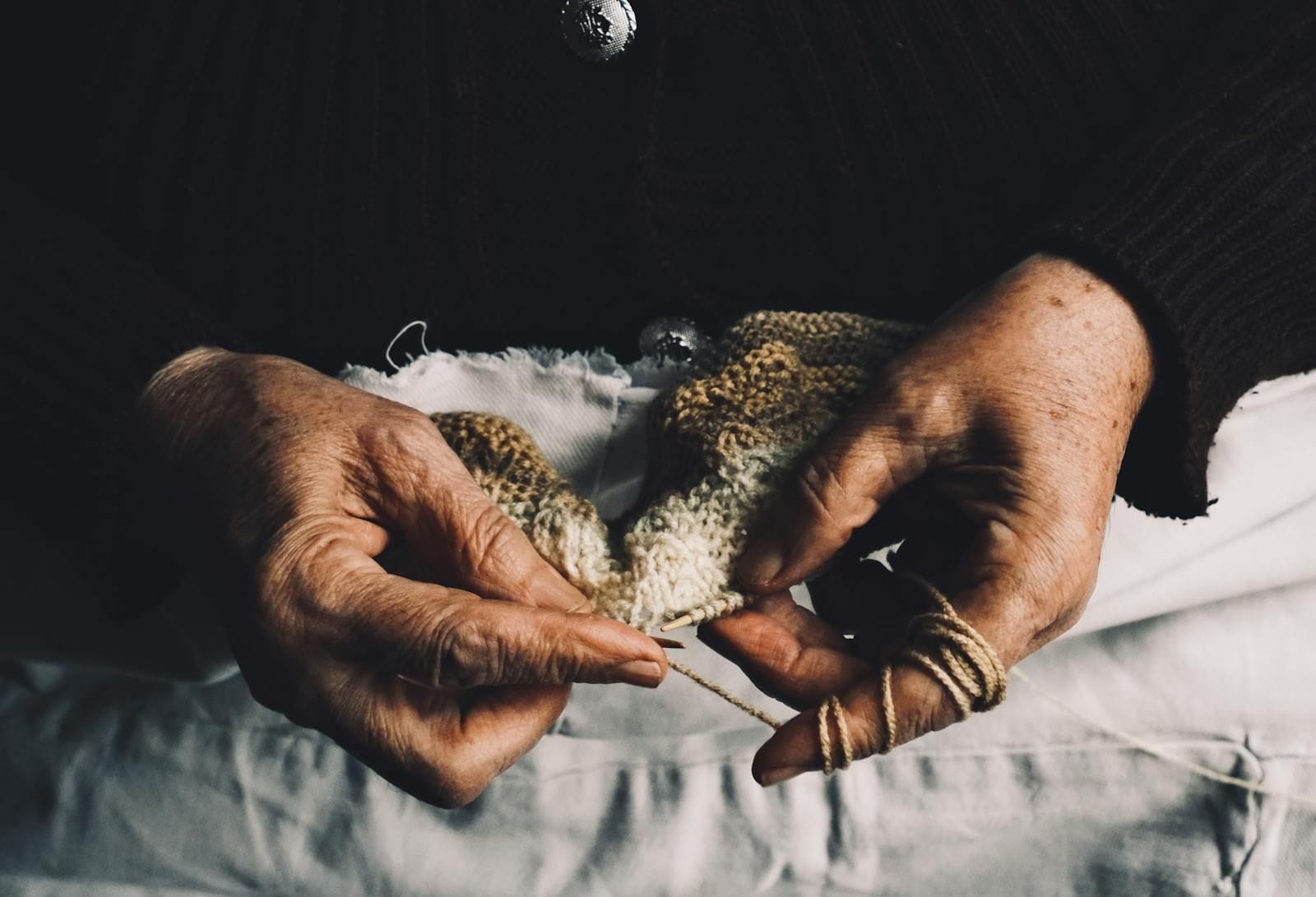 A close-up of an older woman's hands working knitting needles by warm lamplight, the bright wool looped between her fingers, the surrounding scene blurred into a soft glow that emphasizes the careful, practiced motion of the hands themselves