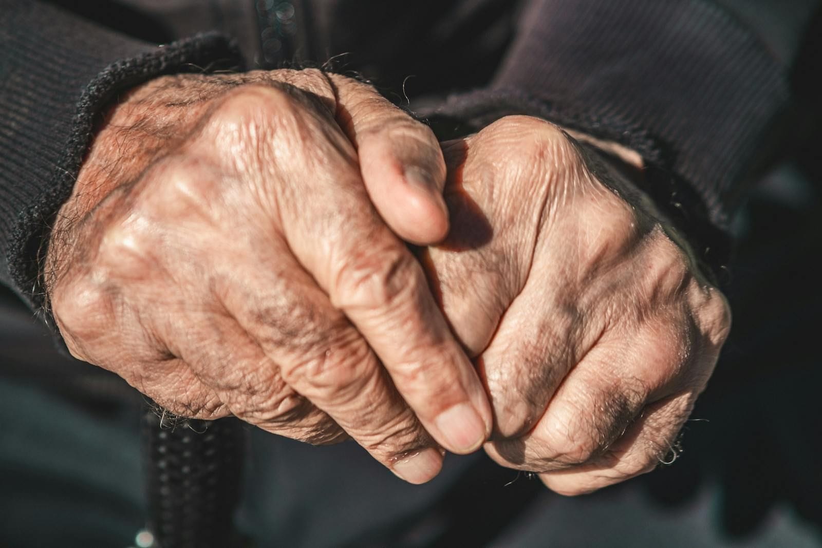 A close-up of a senior person's clasped hands resting in a contemplative pose, the knuckles showing the slight enlargement that years of joint wear leave, the surrounding skin lined and folded, the composition still and quiet