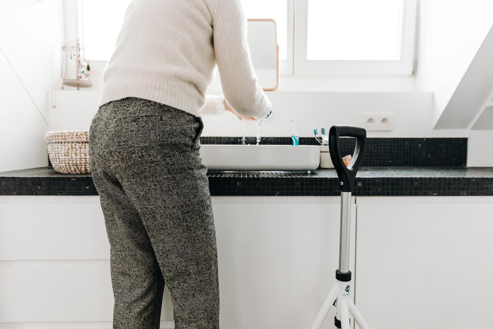 A grey-haired senior in a white sweater and grey trousers moves slowly through a bright modern bathroom alongside a four-wheel rollator, one hand resting on the rollator's frame and the other angled toward a sink under a wide window, the scene reading as ordinary domestic mobility rather than crisis