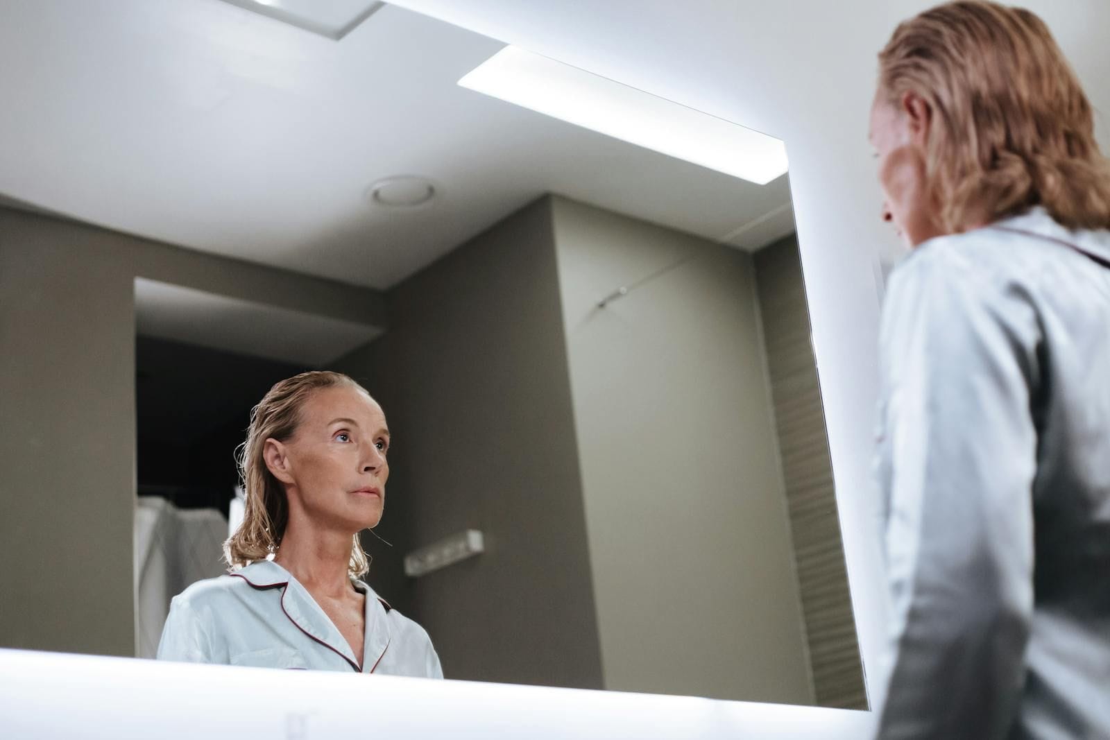 A senior woman with damp dark hair sits looking thoughtfully at her reflection in a softly-lit bathroom mirror, the modern vanity in muted tones around her, the moment reading as the quiet morning recalibration that comes before the rest of the day