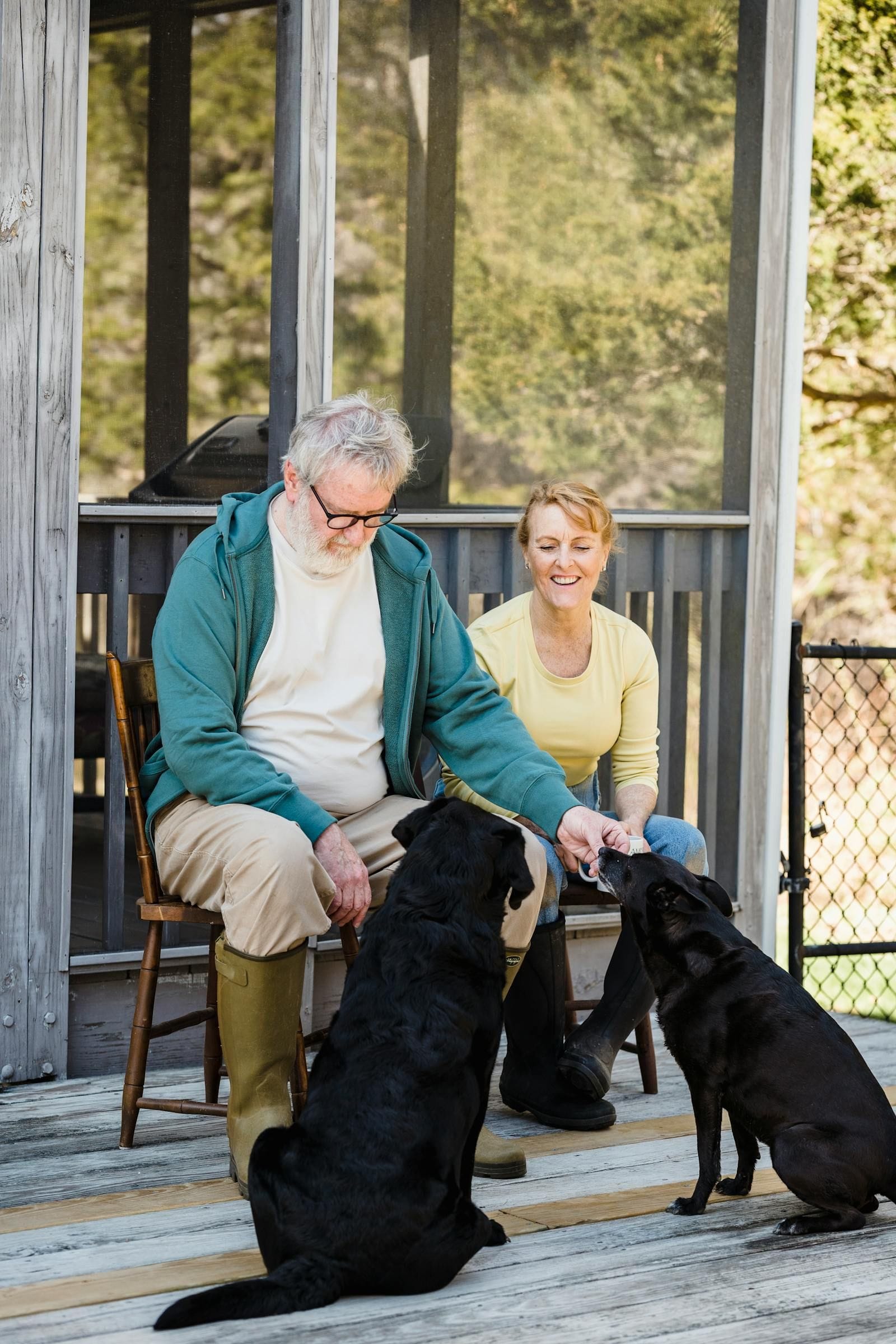 A senior couple sit close together on the wooden steps of a sunlit front porch surrounded by green plants, the silver-haired woman in a coral shirt laughing softly as the white-haired man leans toward her, two calm small dogs at their feet — one tan, one black — looking up between them