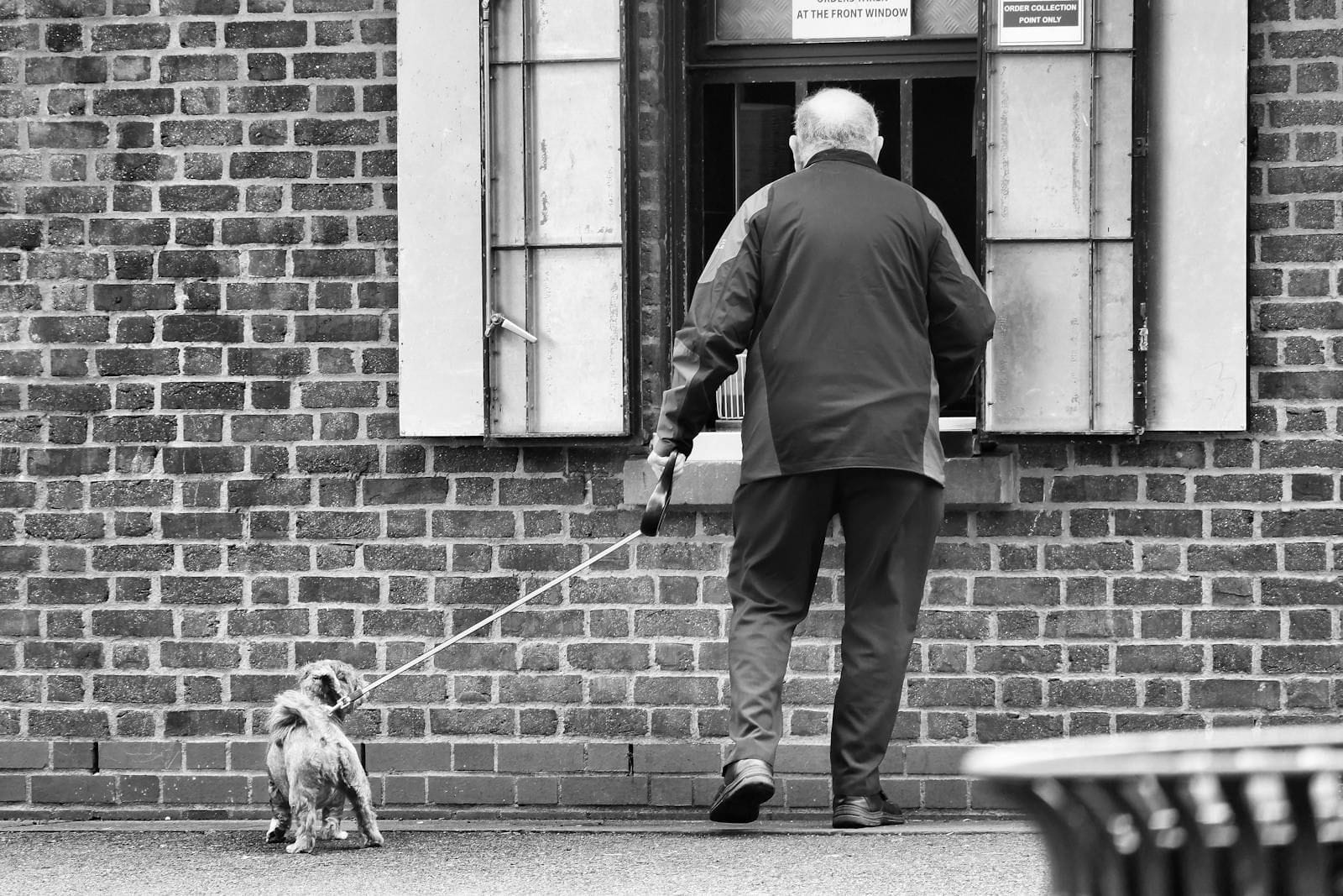 A white-haired older man in a dark grey coat walks slowly along a quiet city sidewalk beside a low brick building, a small light-colored dog on a short leash trotting calmly a step ahead of him, the whole scene caught from behind in the soft grey light of an unhurried morning