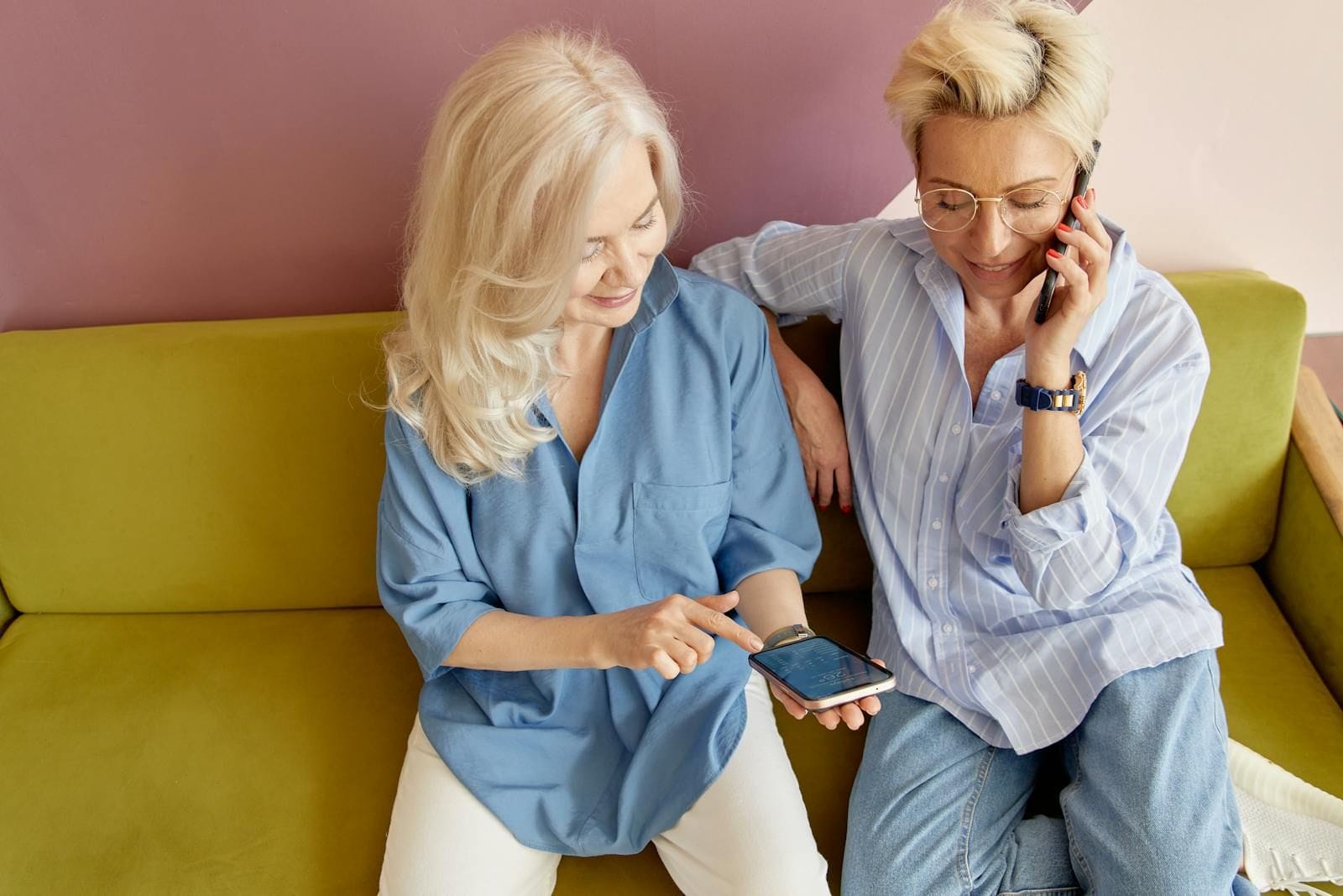 Two silver-haired older women in soft, layered indoor clothing sit close together on a long velvet sofa in a sunlit living room, both leaning toward a single smartphone held between them with the screen tilted upward so they can see it together, an open hardcover book lying face-down on a low coffee table beside a half-empty glass of water