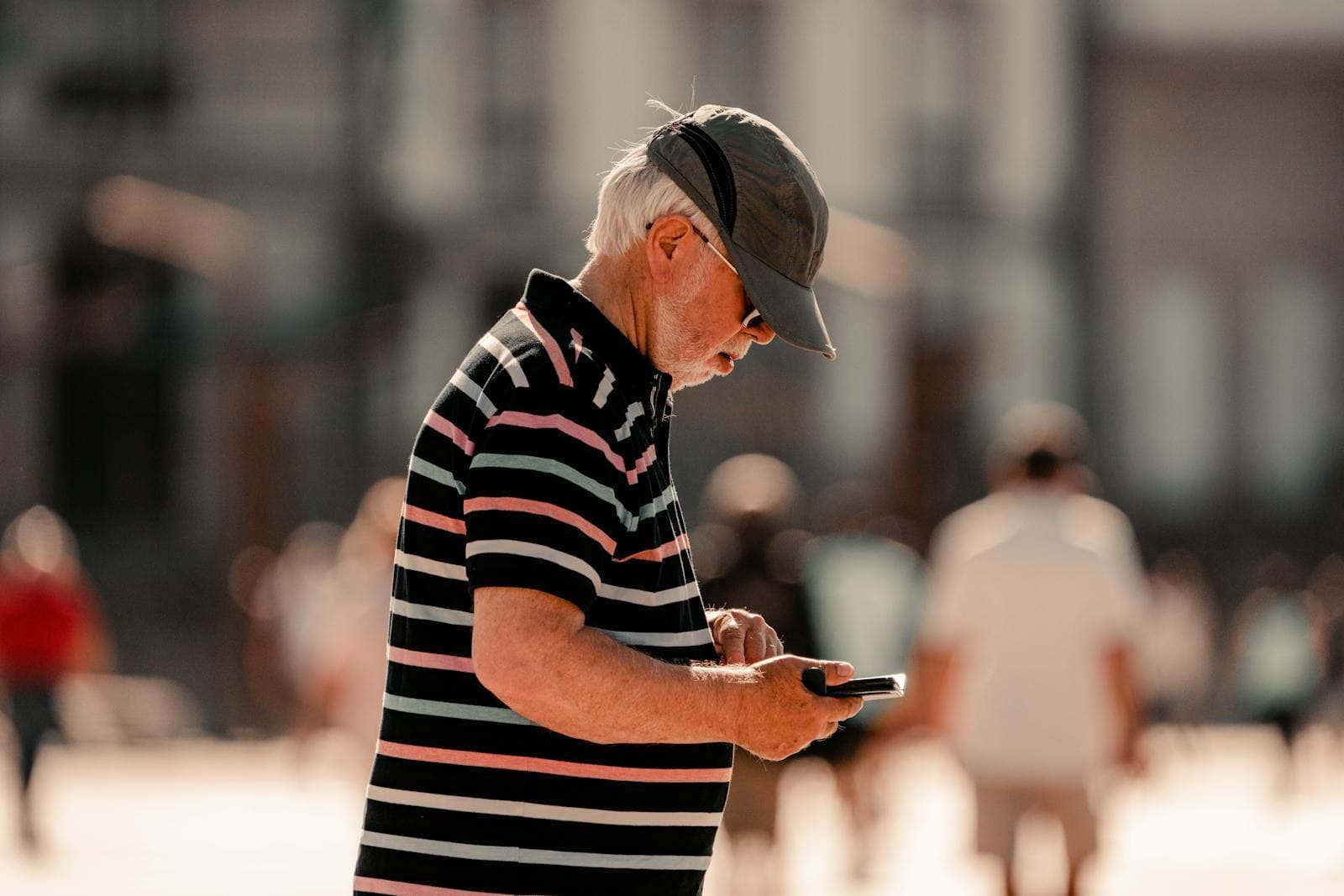 A bearded older man in a vertical-striped pale-blue shirt stands on a sunlit pedestrian street holding a small black smartphone in his right hand and looking down at the screen, the city of La Coruña visible behind him in soft Mediterranean afternoon light with stone-paved sidewalk and warm building facades along the block