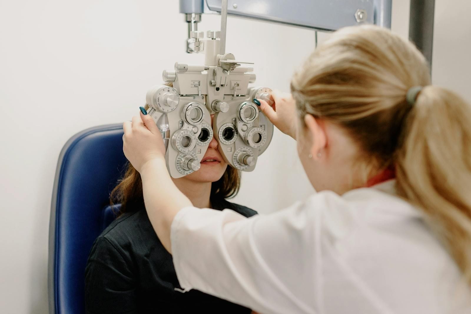 An eye doctor in a bright modern clinic adjusting a phoropter, the multi-lens vision-testing device, in front of a seated patient during a refraction exam