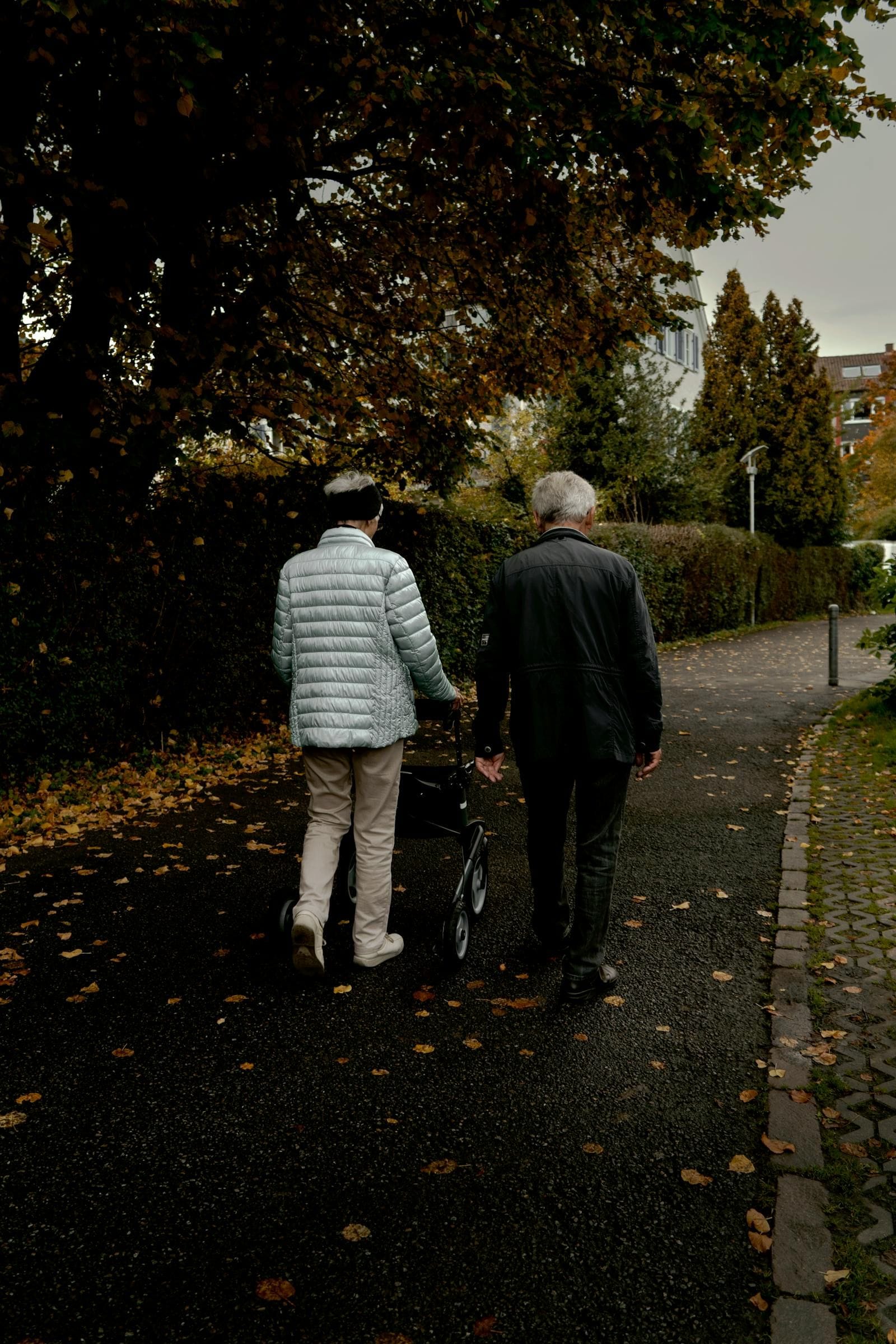 An elderly couple walks together along a leaf-strewn autumn path in a Munich park, one of them using a four-wheeled rollator with the other walking quietly alongside in companionable rhythm