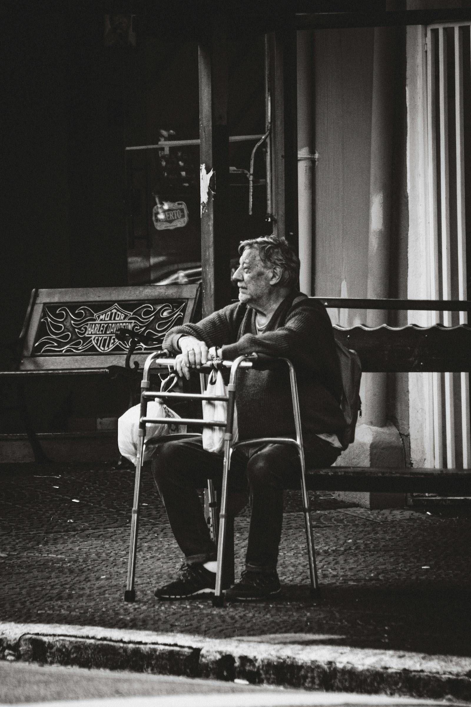A senior man sits at rest on the built-in seat of his rollator on a city sidewalk in Sao Paulo, hands resting on the grips, taking the kind of brief pause the seat exists to make possible
