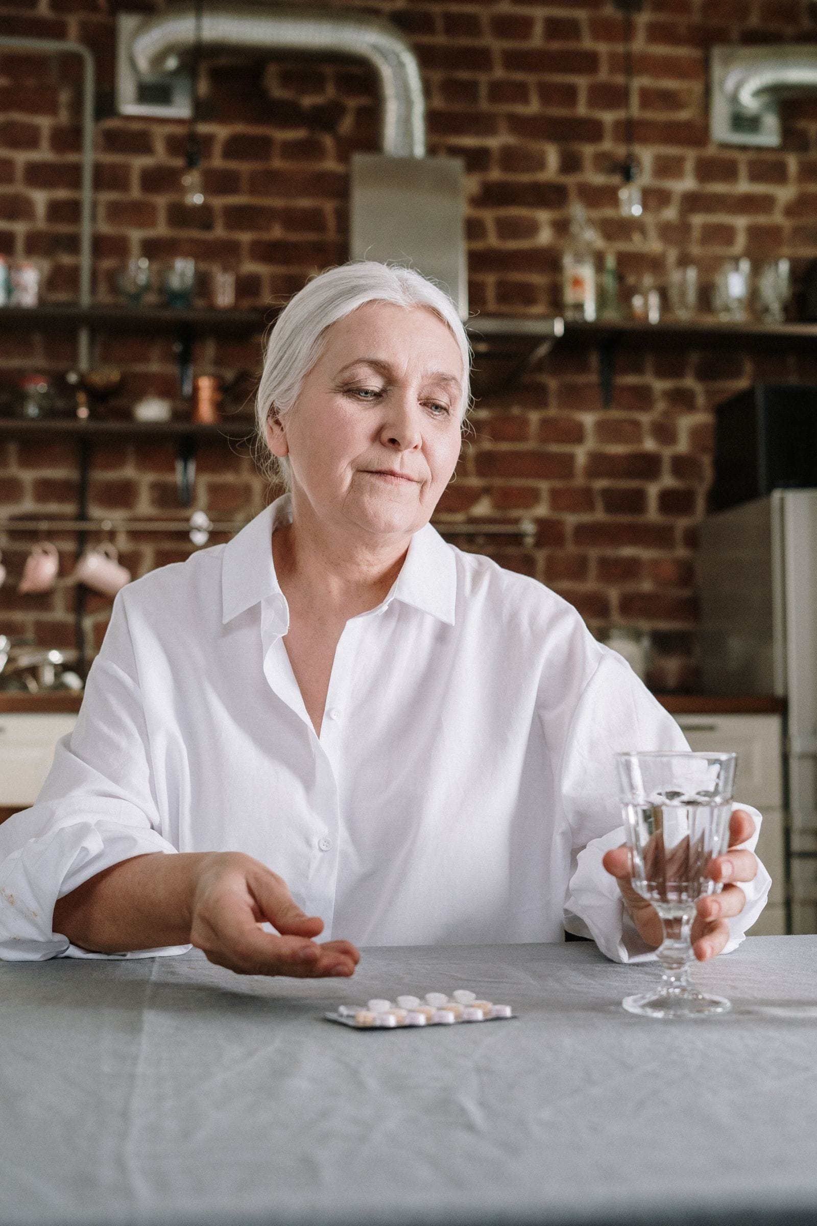An older woman sitting at a kitchen table holding a pill in one hand and a glass of water in the other