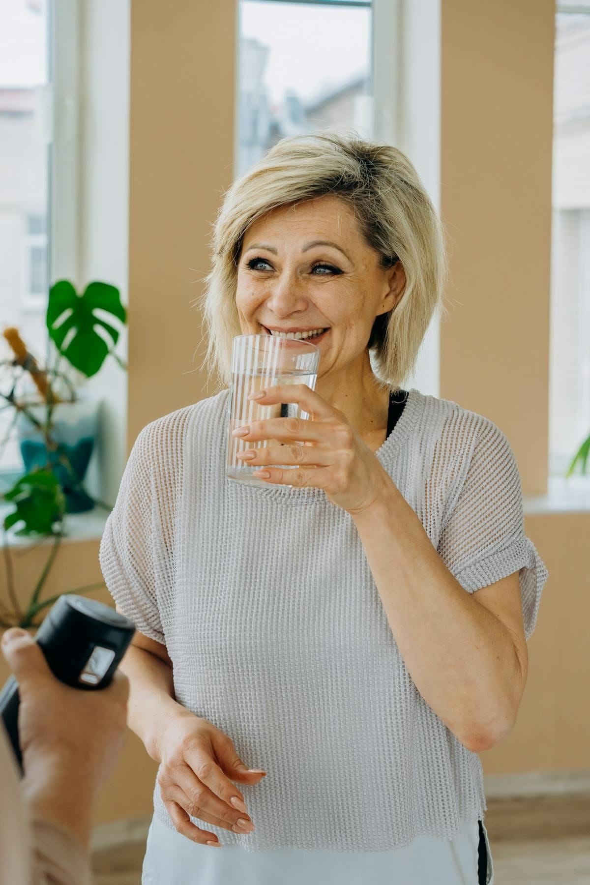 A smiling silver-haired woman in a bright, plant-filled living room raising a glass of water to take a drink