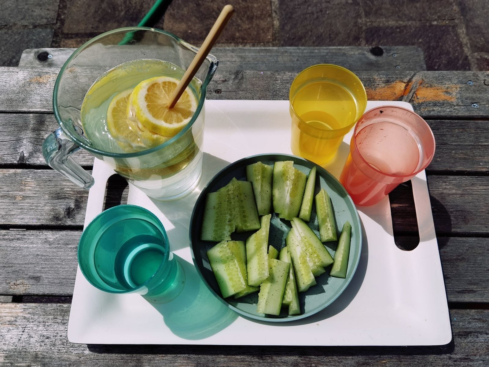 An outdoor wooden table set with a glass of lemon-infused water, a small pitcher, and fresh cucumber slices on a sunny day
