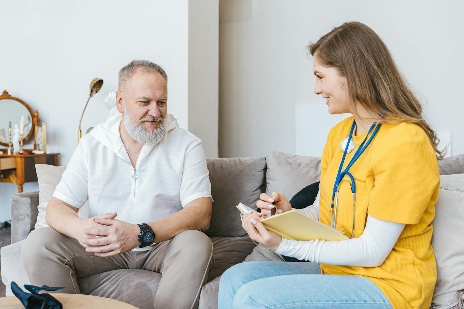 A healthcare professional in scrubs sits beside an elderly man on a sofa with a stethoscope around her neck, leaning forward with a warm, attentive expression as she talks him through a question, illustrating the kind of careful, unhurried medical evaluation that brings the cause of a sudden confusion to light
