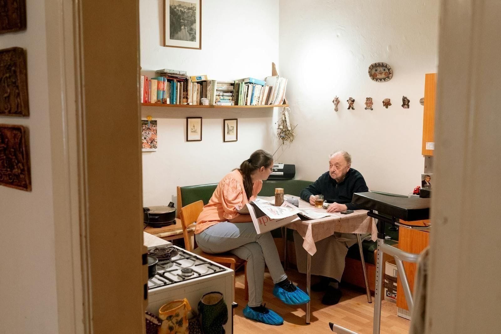 An older woman and a caregiver sit close together at a small kitchen table with steaming cups of coffee between them, the caregiver leaning in with a soft attentive smile while the older woman talks, capturing the quiet, unhurried company that does much of the work of recovery from delirium