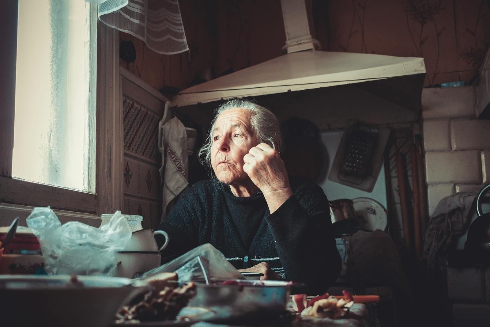 An older woman sits at a rustic wooden kitchen table beside a window, gazing out into the soft daylight with a quiet, faraway expression, hands resting in her lap as if watching for something only she can see