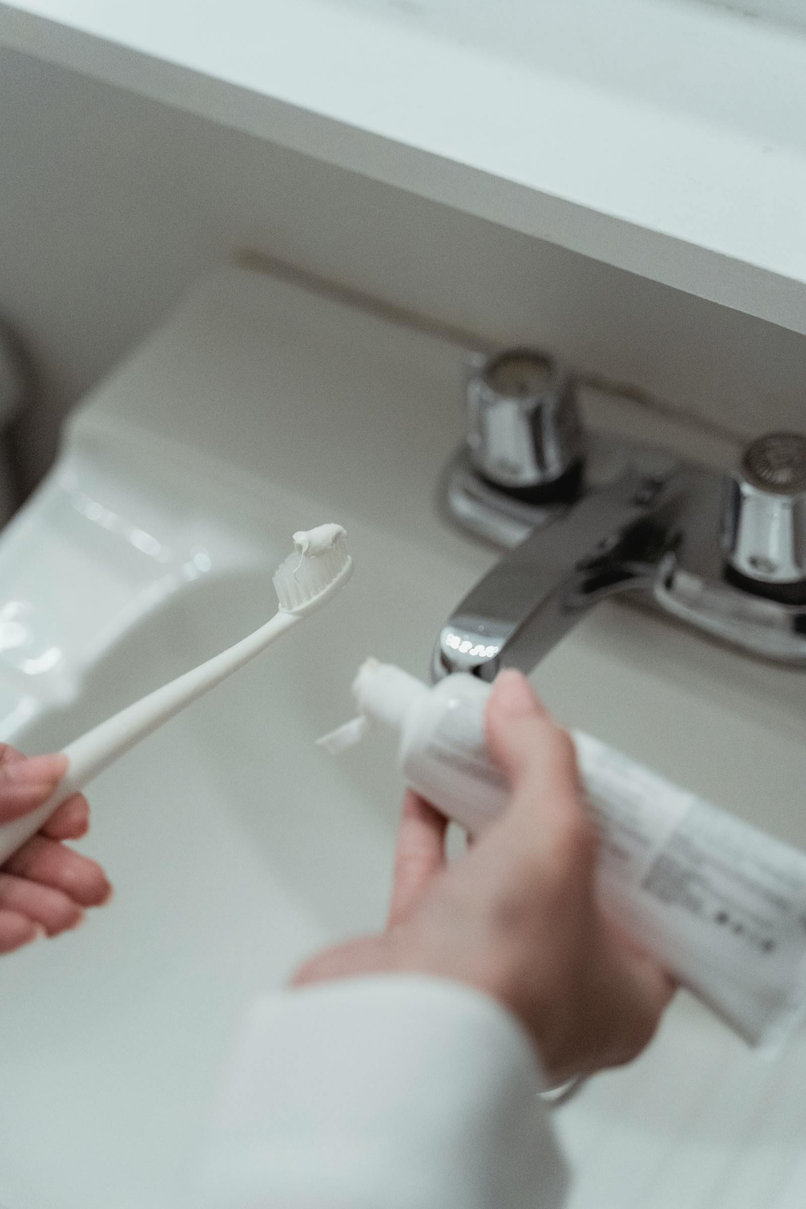 A close-up of a hand squeezing toothpaste onto a toothbrush over a clean white bathroom sink