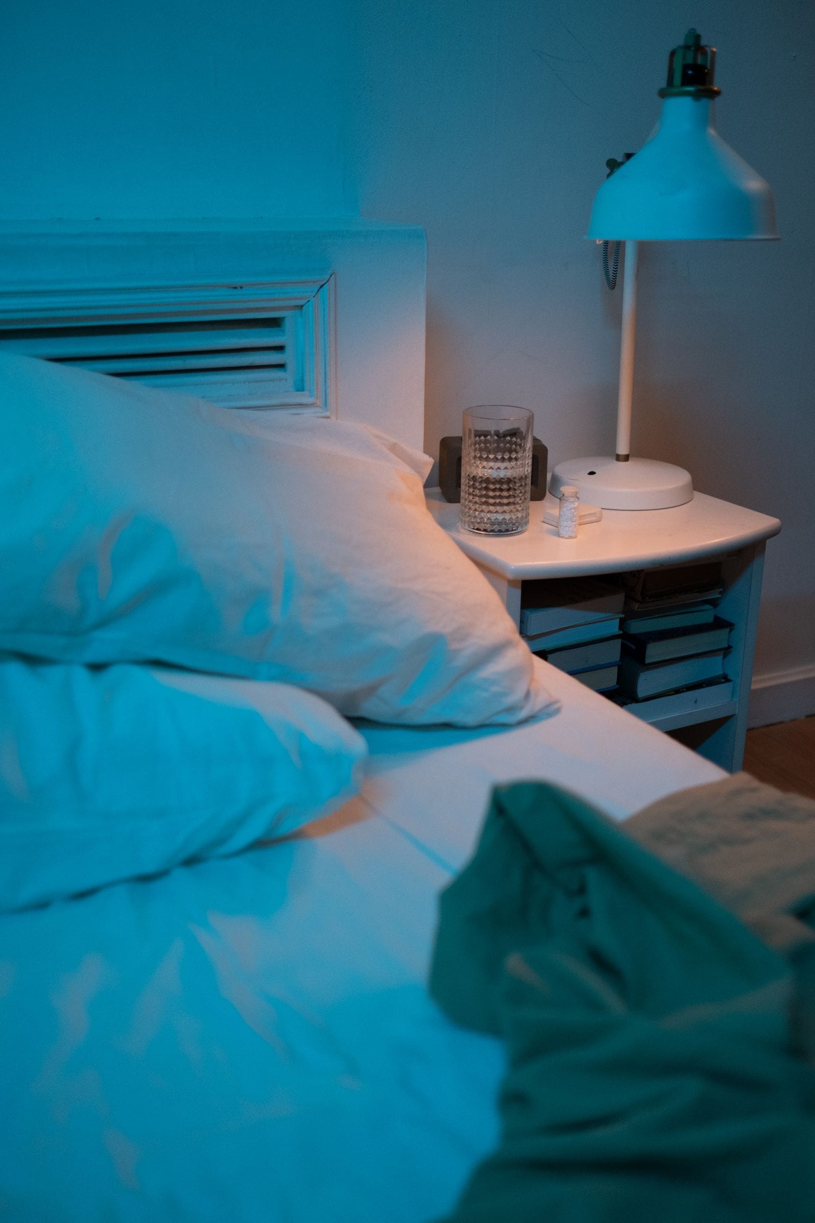 A glass of water and a small bottle of pills on a bedside table beside a lamp at night, next to a bed
