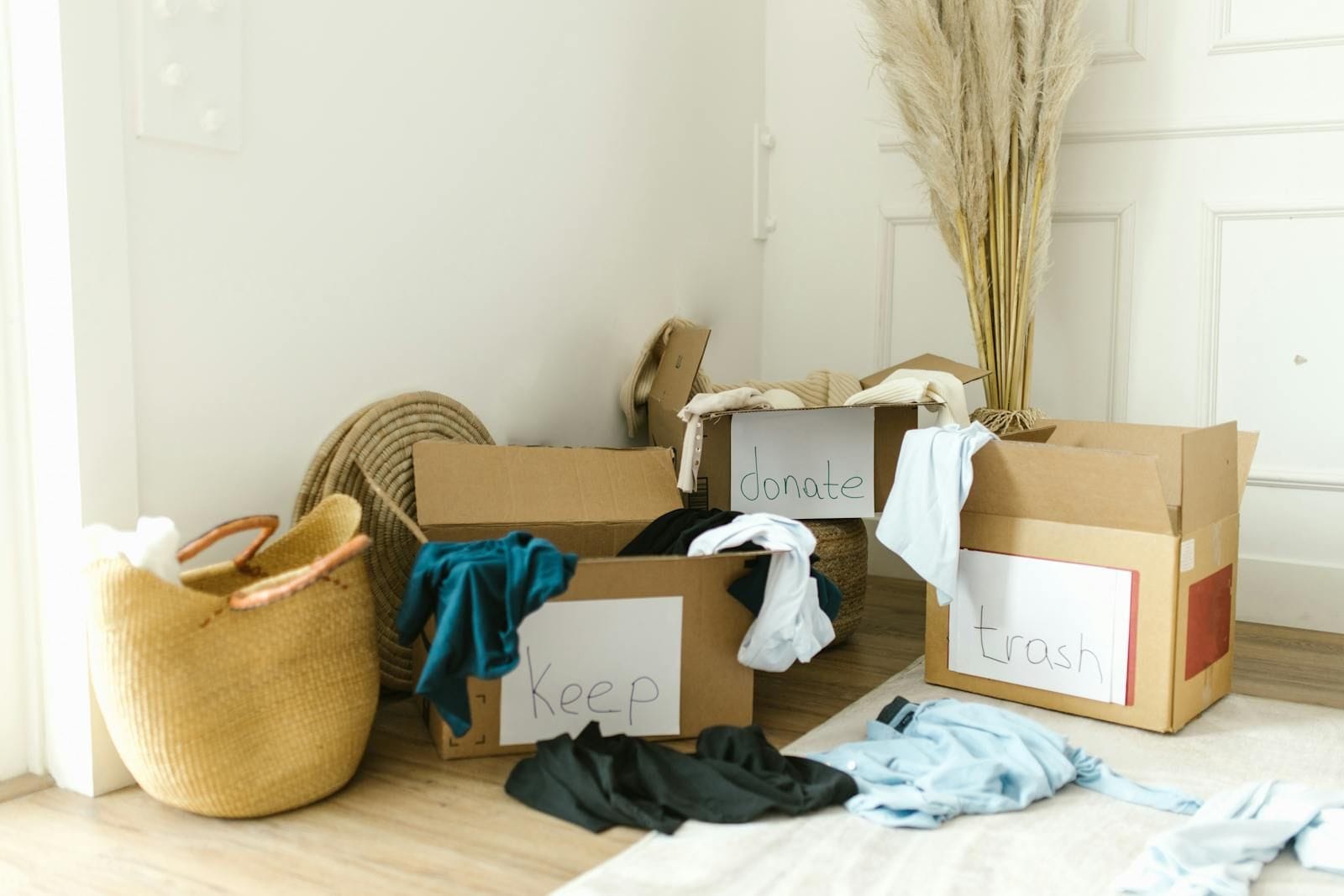 Three open cardboard boxes on a wood floor labeled by hand keep, donate, and trash, with folded clothing sorted between them and a woven basket alongside, illustrating a simple sorting system