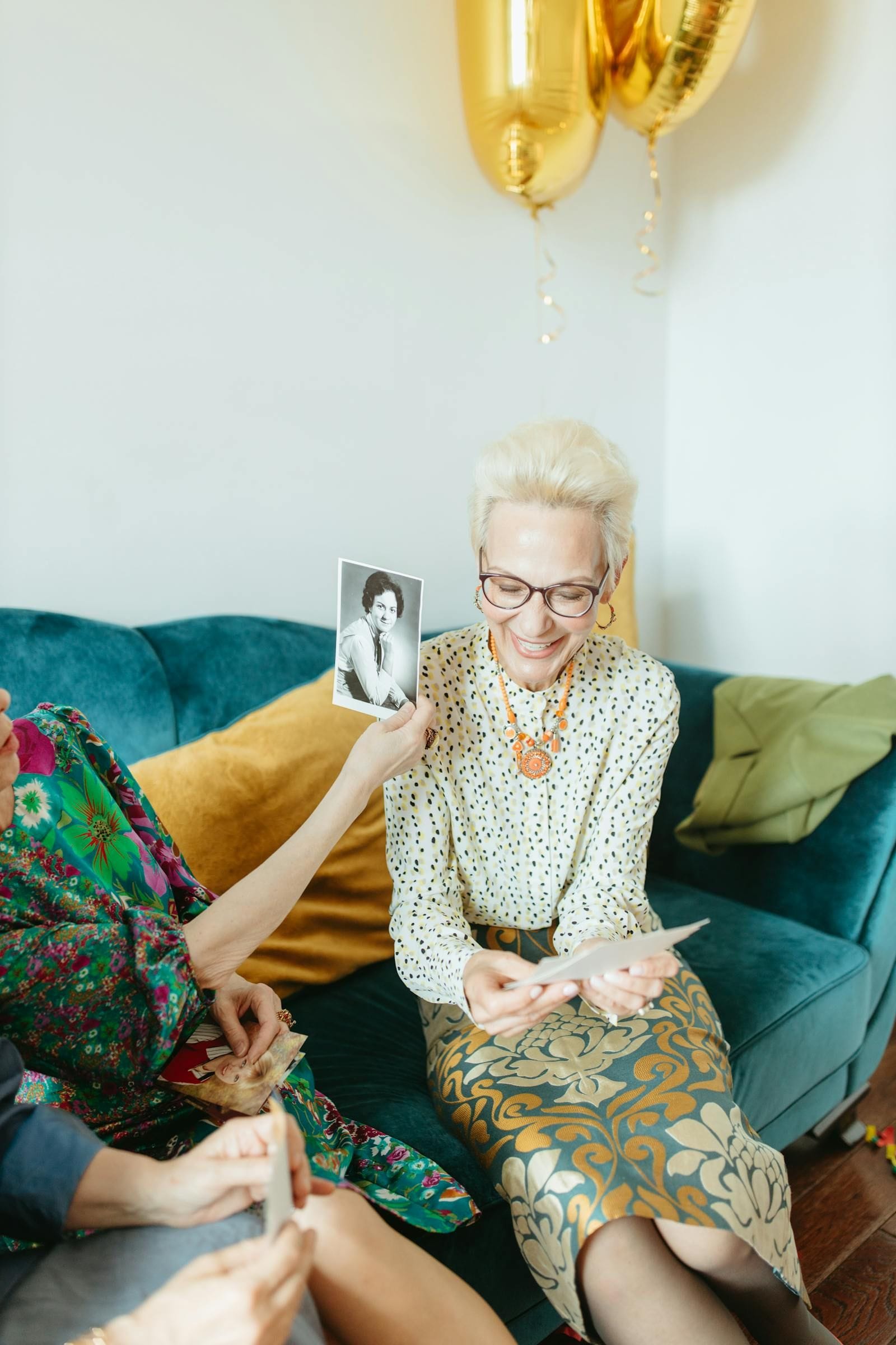 A smiling older woman in glasses and a polka-dot blouse holding up a vintage black-and-white portrait photograph while sitting on a teal velvet sofa, sharing keepsakes with a companion seated beside her
