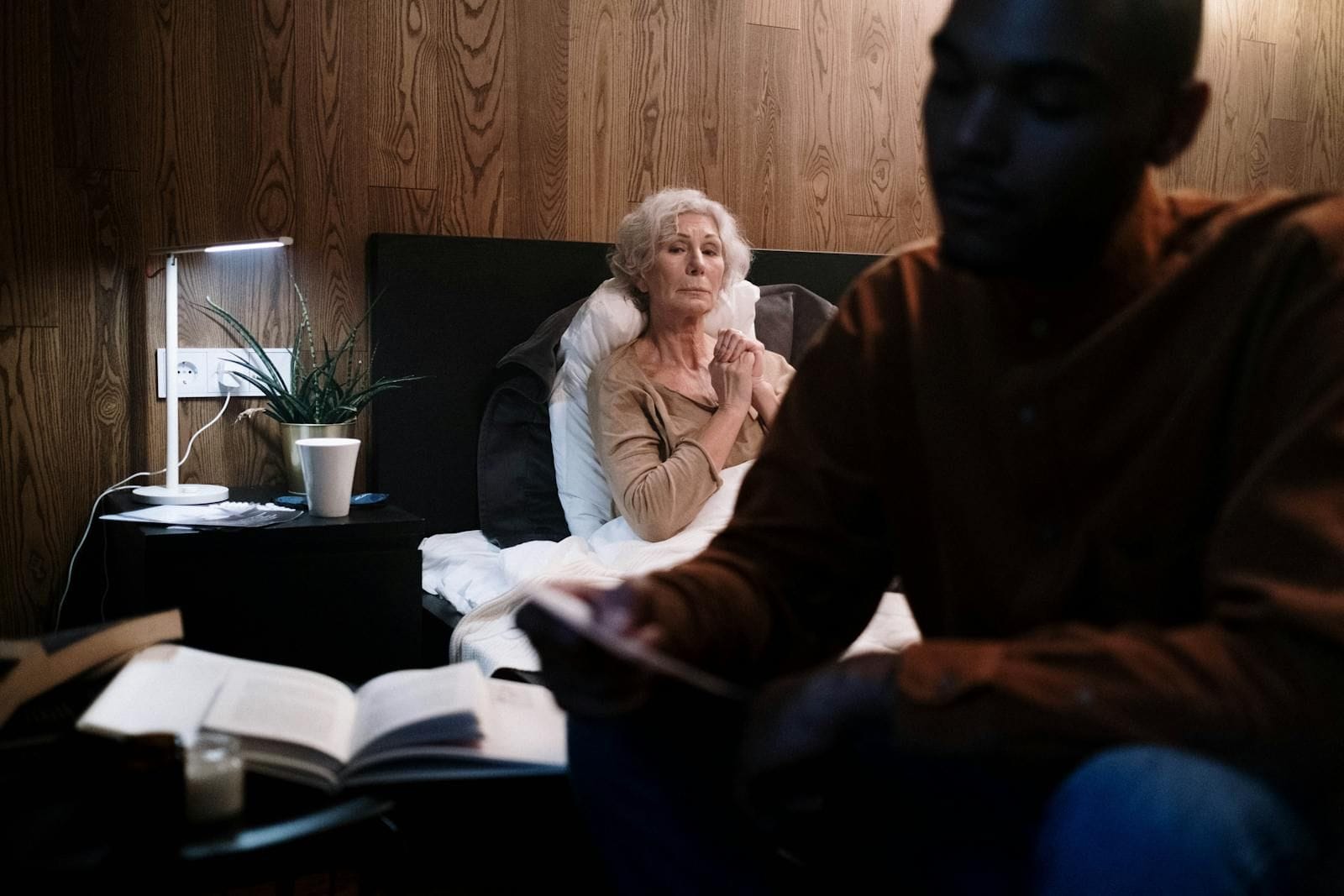 A grown grandson sits on the edge of a bed in the warm gold of a single bedside lamp, leaning forward and looking down at his grandmother resting under the covers, the bedroom around them quiet and softened by evening light, the unhurried presence of someone who has decided this is where he needs to be tonight