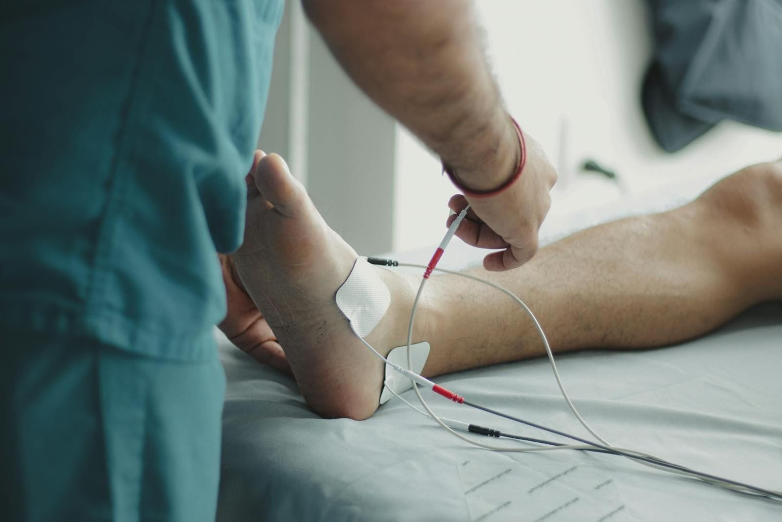 A clinician in dark teal scrubs stands beside an exam table and gently steadies a patient's bare foot with one hand while connecting a small wired sensor along the ankle for a nerve-function check, the foot pale against the soft hospital sheets and the wires running back toward an unseen monitor at the head of the bed