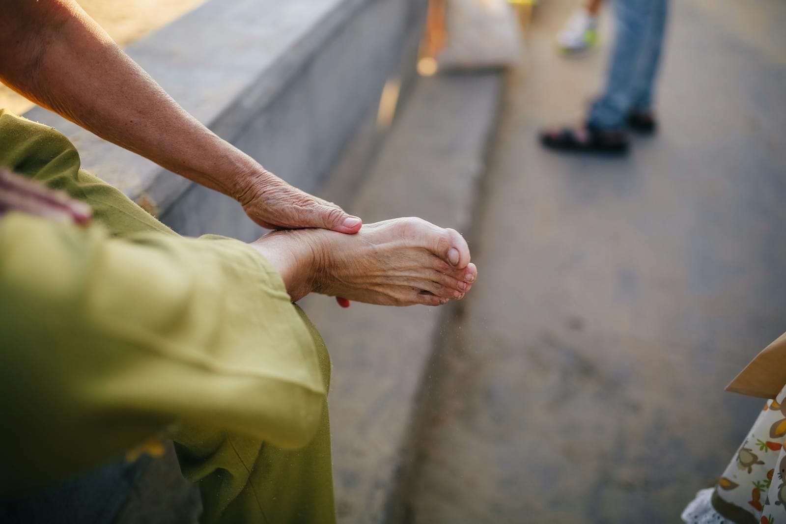 An older woman in an olive-green dress sits outdoors on warm stone steps with one bare foot resting on her knee, her aged hand wrapped gently around the foot in the kind of unhurried self-inspection that catches a small problem before it becomes a large one, the late afternoon light catching the lined skin of both her arm and her foot
