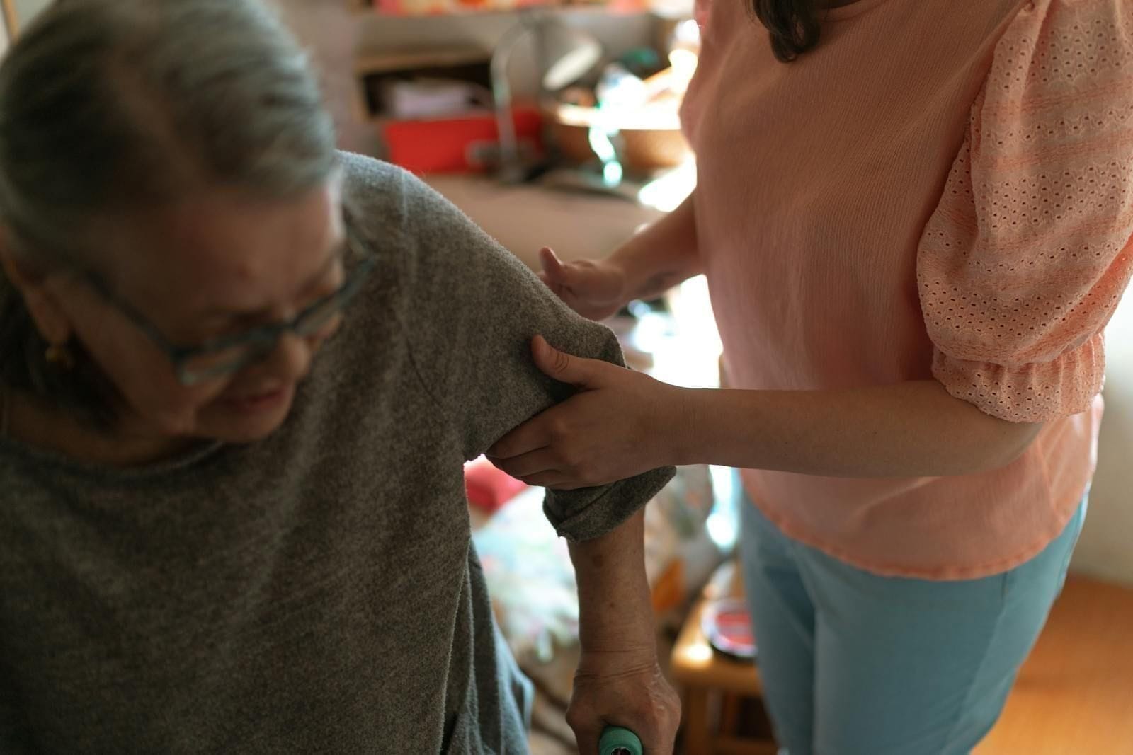 A home caregiver in a soft beige sweater leans in to help an elderly woman sit up safely in her bedroom, a four-wheeled walker positioned just beside the bed, the room lit by warm afternoon window light filtering through linen curtains