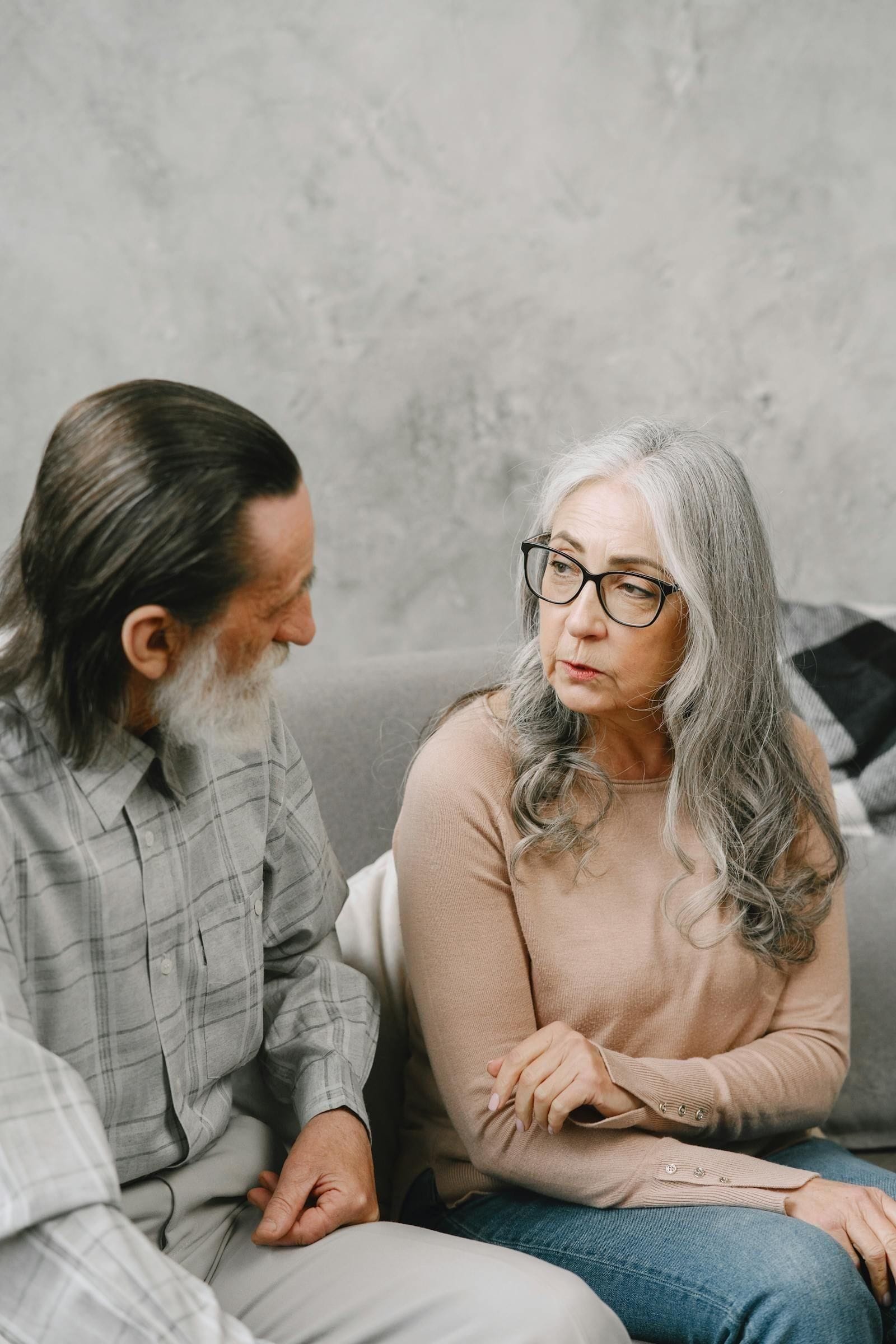 An older couple sit close on a couch in soft gray light, the husband leaning toward his wife in mid-sentence and the wife looking down with her hands folded, illustrating the kind of slow, heavy conversation families have when the question of palliative care or hospice first comes up