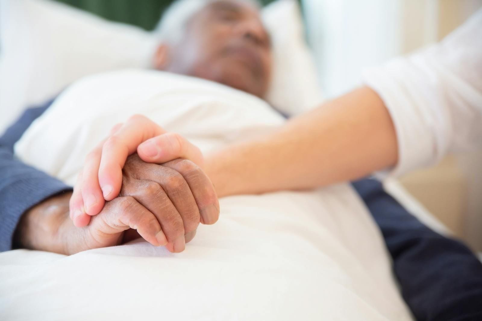 A close-up of two pairs of hands clasped together on the white linens of a bed, one set wrinkled and resting on the patient's chest, the other belonging to a younger family member or caregiver bent close in support
