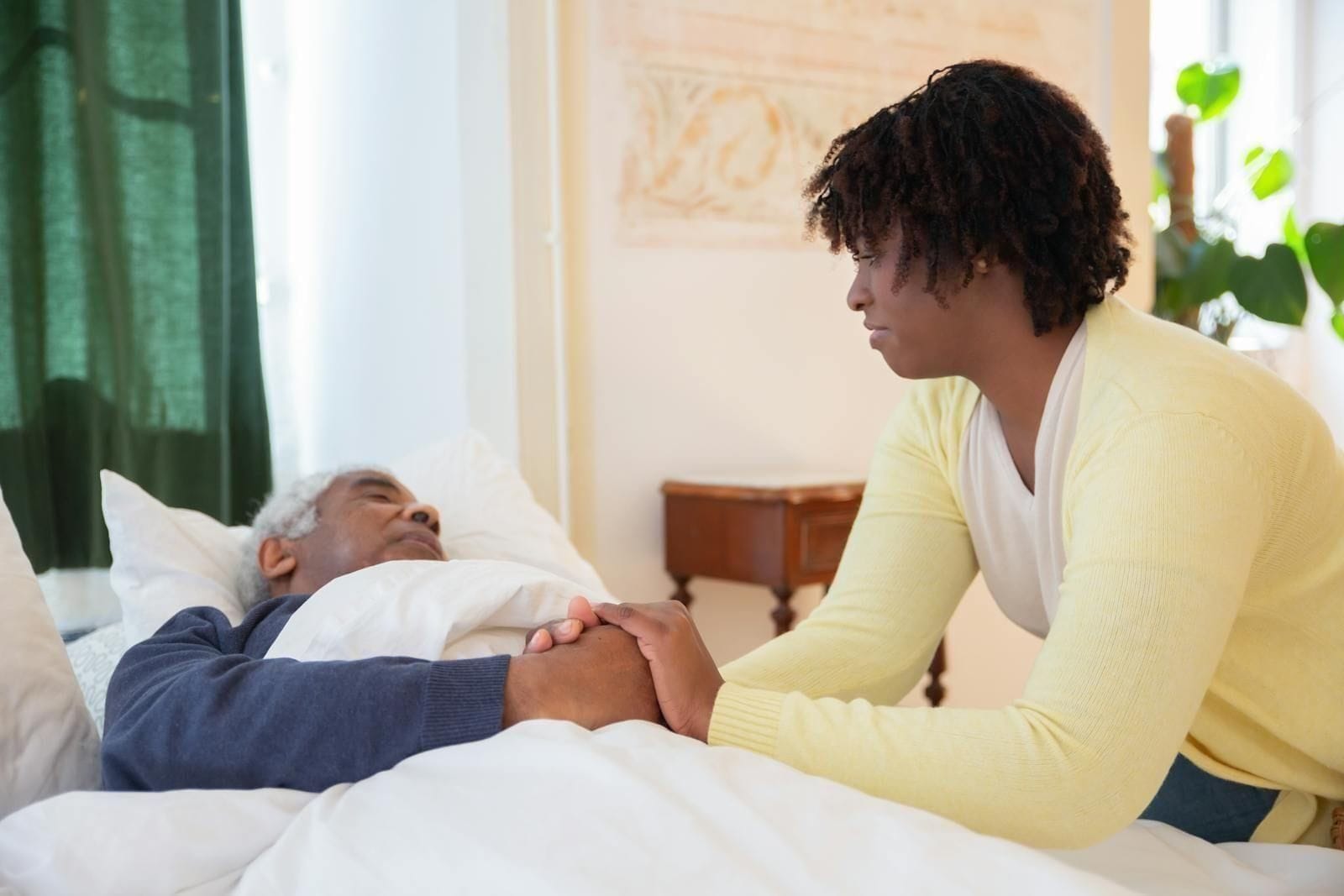 A caregiver in a soft yellow cardigan leans over a sunlit bed and holds the hand of an older man resting on white linens, the room bright and unhurried around them