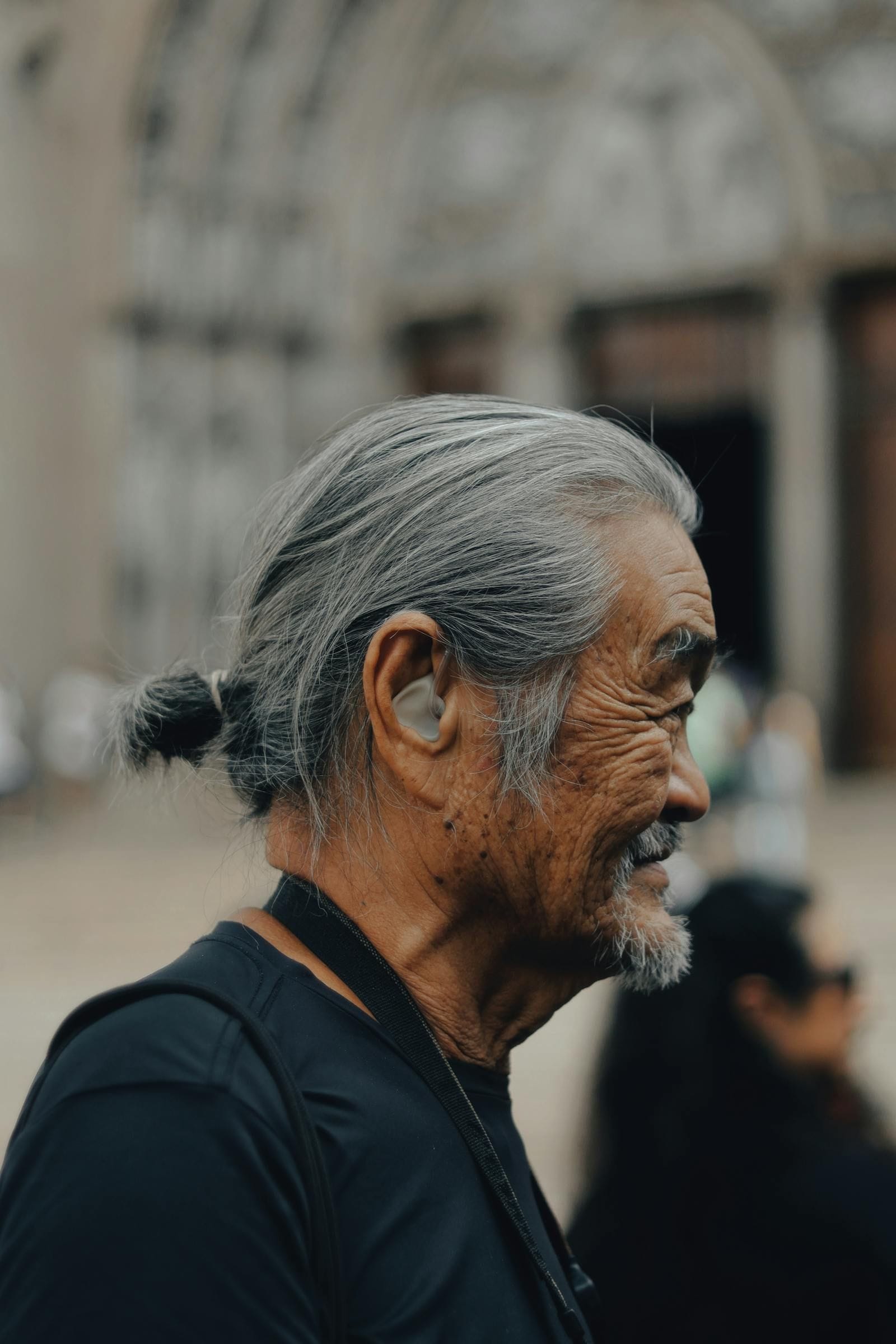 A side-profile portrait of an older man with closely cropped silver hair photographed in soft outdoor light, a small beige behind-the-ear hearing aid visible behind his ear, his expression relaxed and looking off into the middle distance