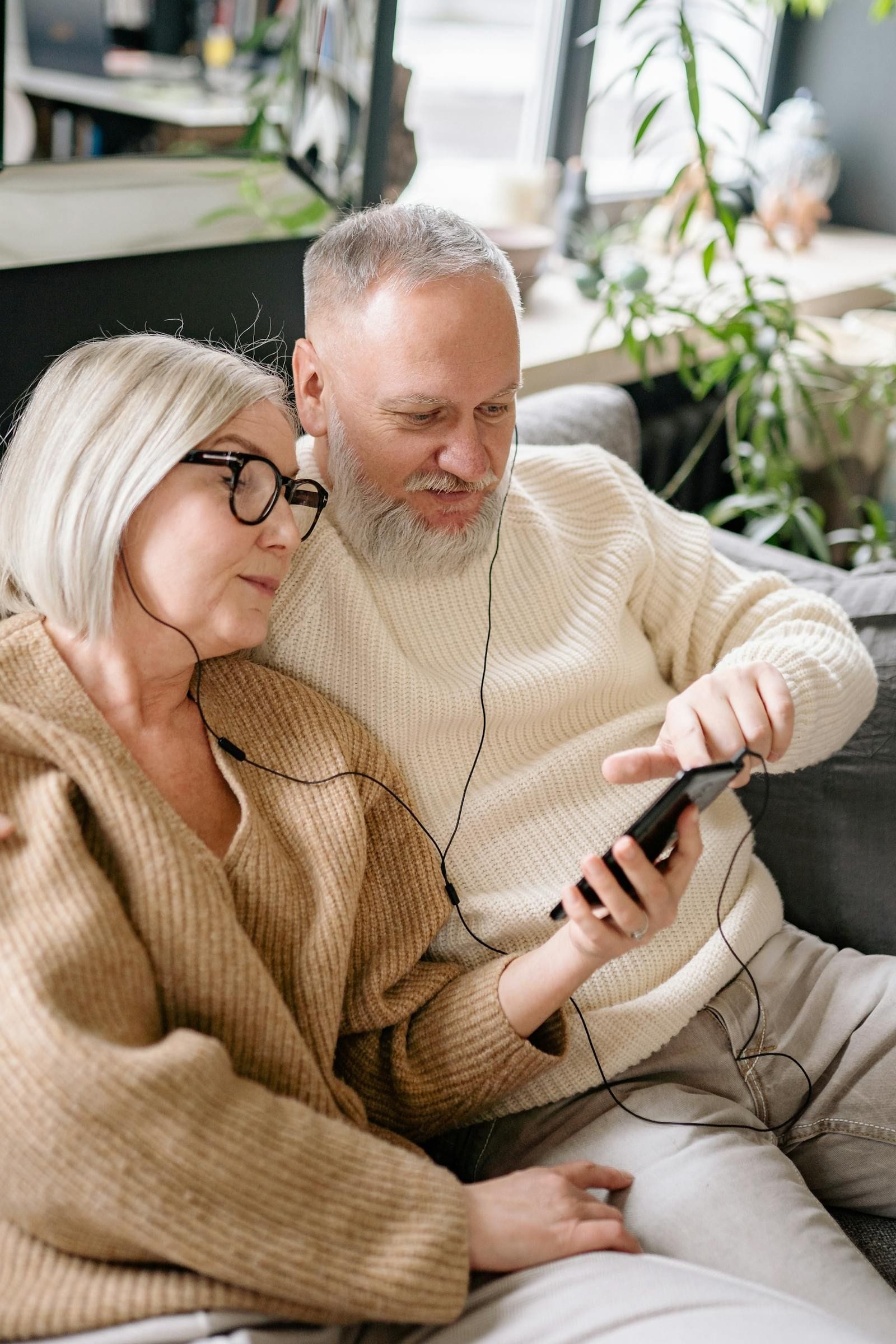An older couple in knitted cardigans sits closely together on a softly lit indoor sofa, the woman leaning in to share a pair of white wired earbuds with the man, both faces relaxed and attentive as they listen to something on a smartphone resting between them