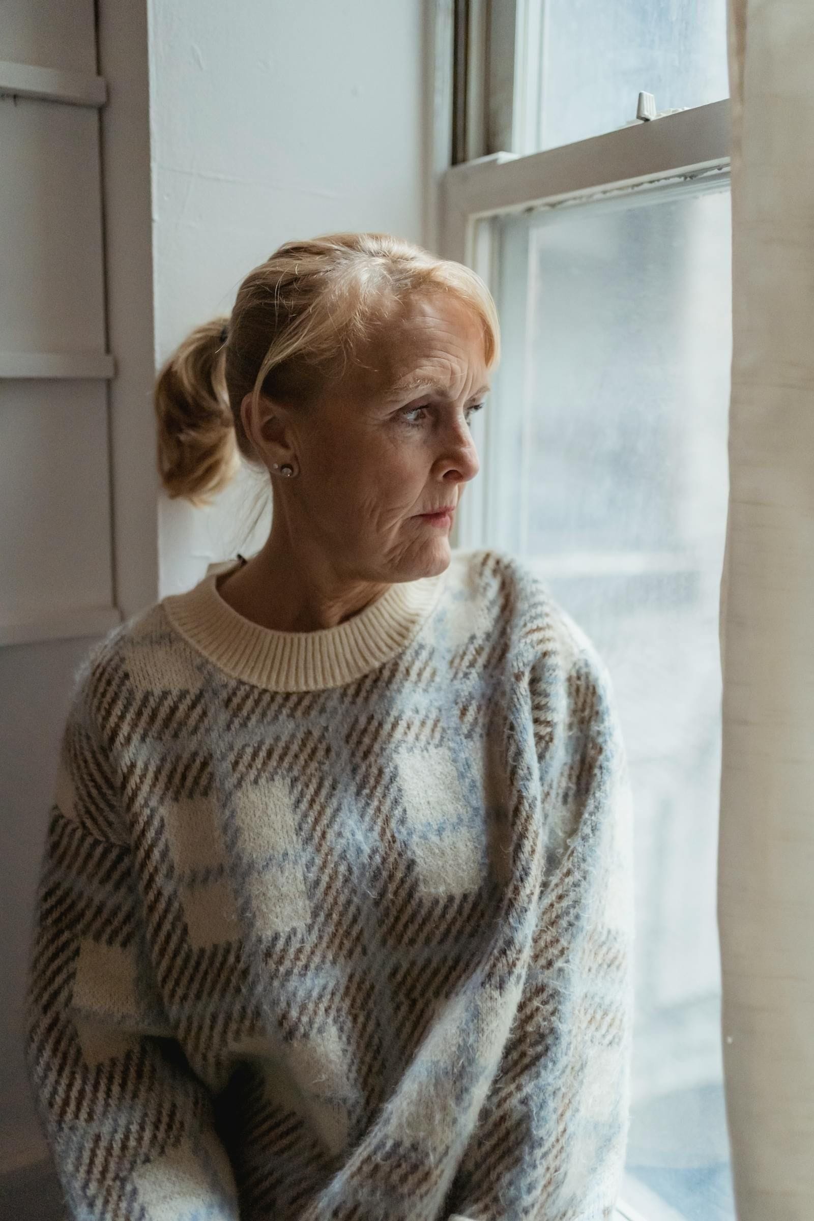 A mature woman in a warm beige sweater sits on a sunlit windowsill at home, knees drawn up, gazing out the window with a quiet, faraway expression that captures the still pause of a caregiver between the demands of one task and the next