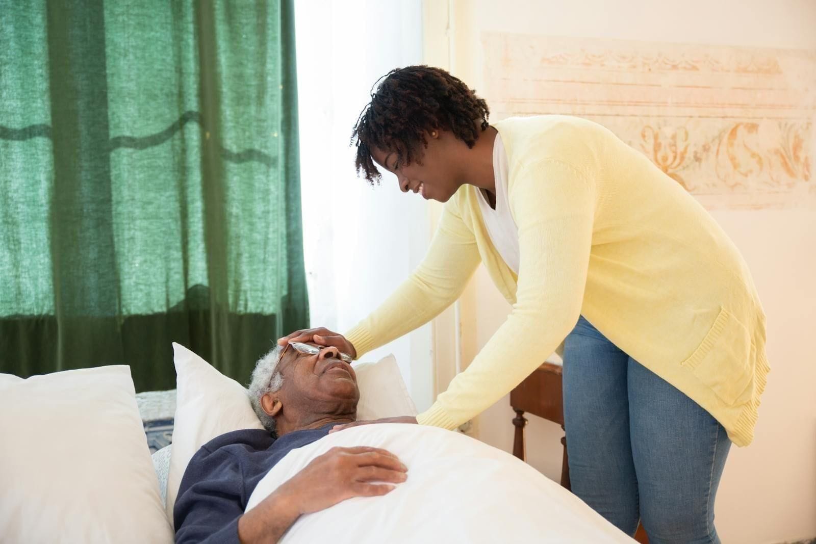 An adult daughter in a soft sweater leans close to her elderly father resting in bed, one hand cradling the back of his head while the other rests against his cheek, the bedroom warm and bright around them in a moment of intimate daily care