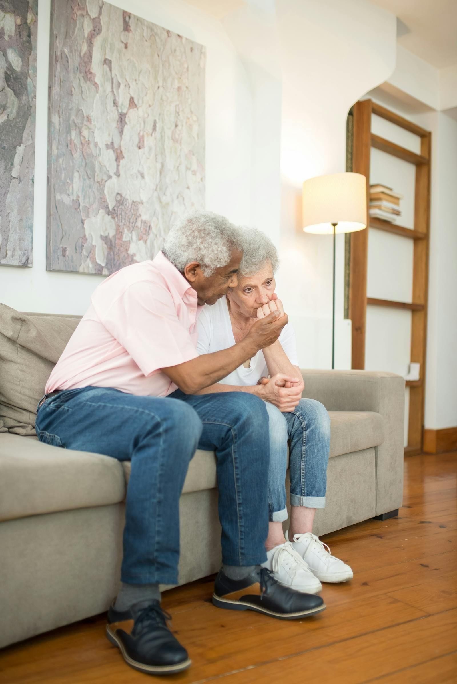 An older couple sit close together on a sunlit couch at home, the wife in a soft pink cardigan leaning her head against her husband's shoulder, his arm wrapped quietly around her as they share a long, wordless pause in the day