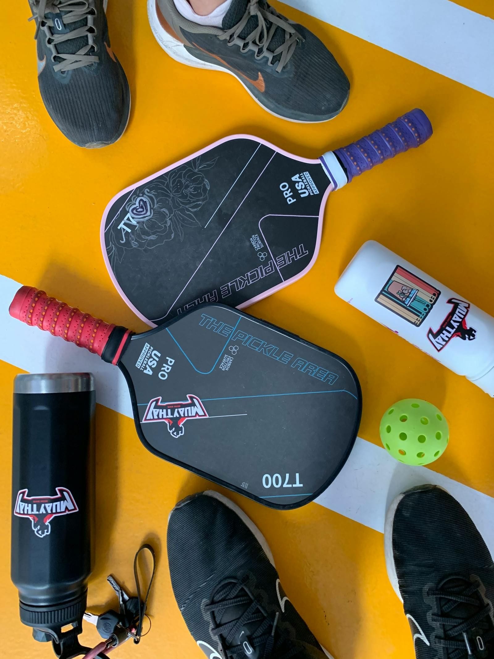 A flat-lay photograph viewed from directly above shows two carbon-fiber pickleball paddles in dark and indigo grips arranged diagonally on a bright yellow concrete court, a yellow pickleball with circular drilled holes resting between them, a black insulated water bottle and two pairs of court shoes framing the corners of the composition