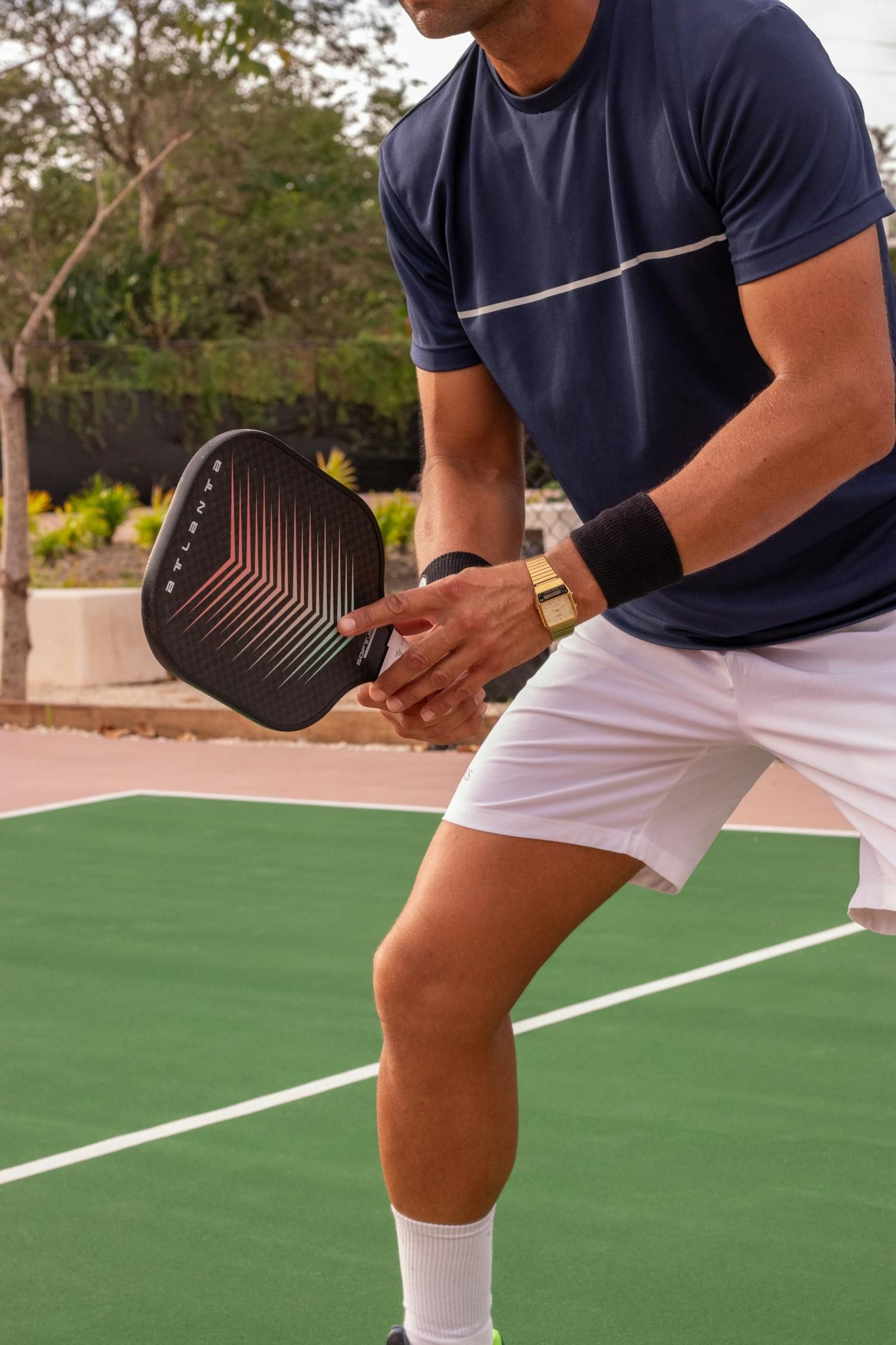 A trim athletic man in a navy short-sleeved shirt and white shorts crouches low in the ready position on a green outdoor pickleball court, his right hand cradling a black carbon-fiber paddle by its face while his left arm braces forward, both knees bent and his weight balanced over the balls of his feet just behind a crisp white sideline