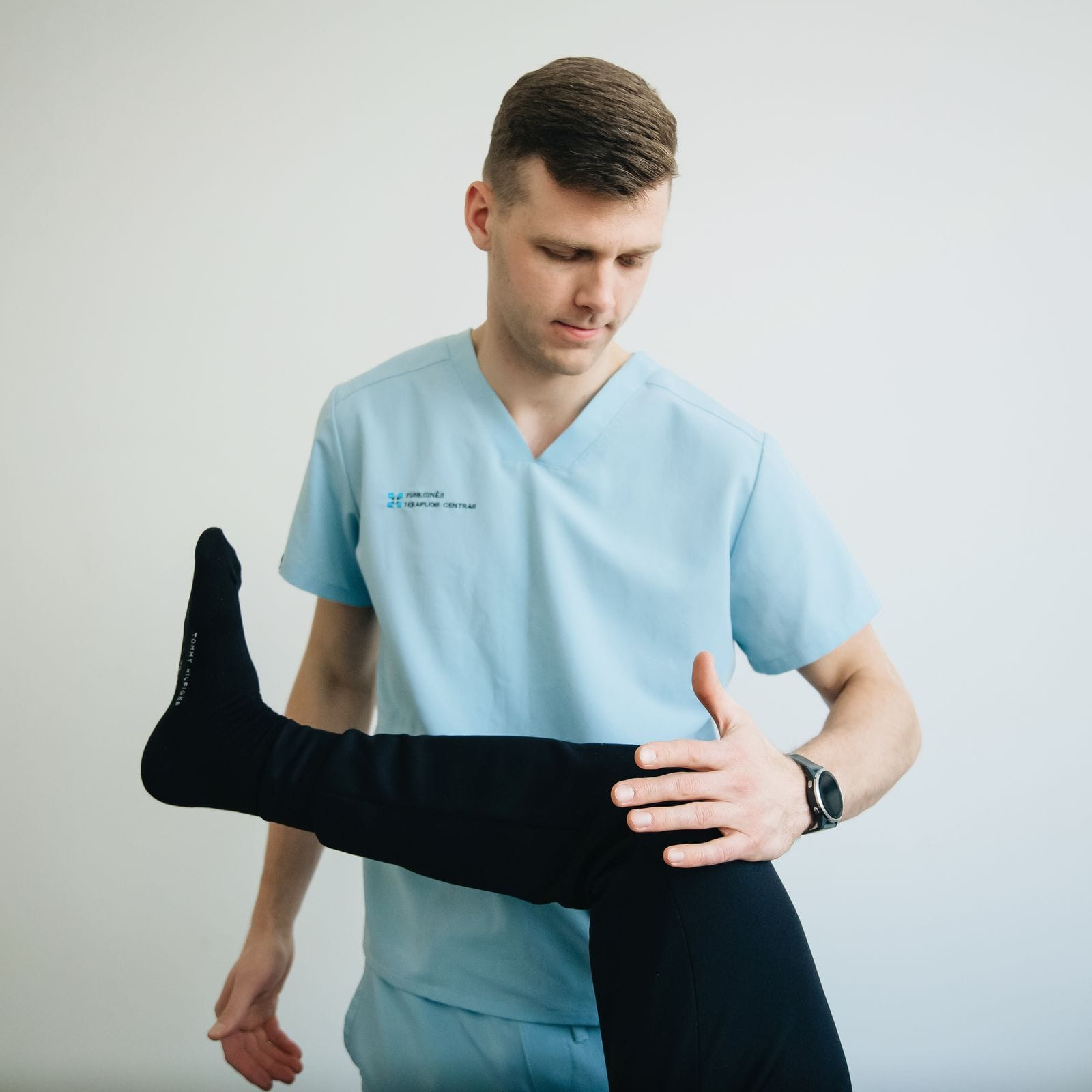 A young male physiotherapist in light blue clinical scrubs with a small clinic logo on the chest standing beside a treatment table and using his right hand to support a patient's lifted left leg by the knee, the patient wearing black sweatpants and a black Tommy Hilfiger athletic sock, the background a soft pale studio wall