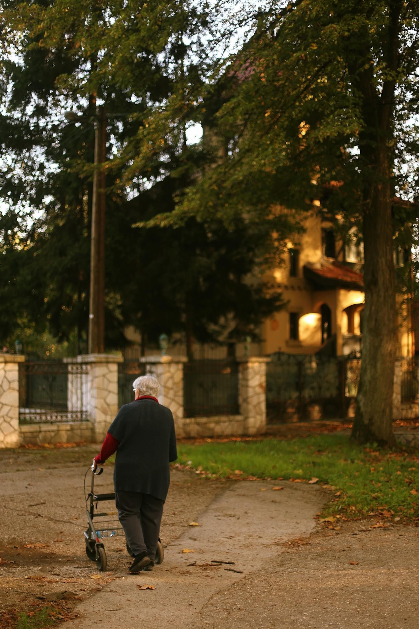 An older woman with white hair wearing a dark coat and dark trousers walking away from the camera down a leaf-strewn paved path beside a low stone garden wall toward a warmly lit stucco villa under autumn trees at dusk, pushing a four-wheeled rollator walker ahead of her