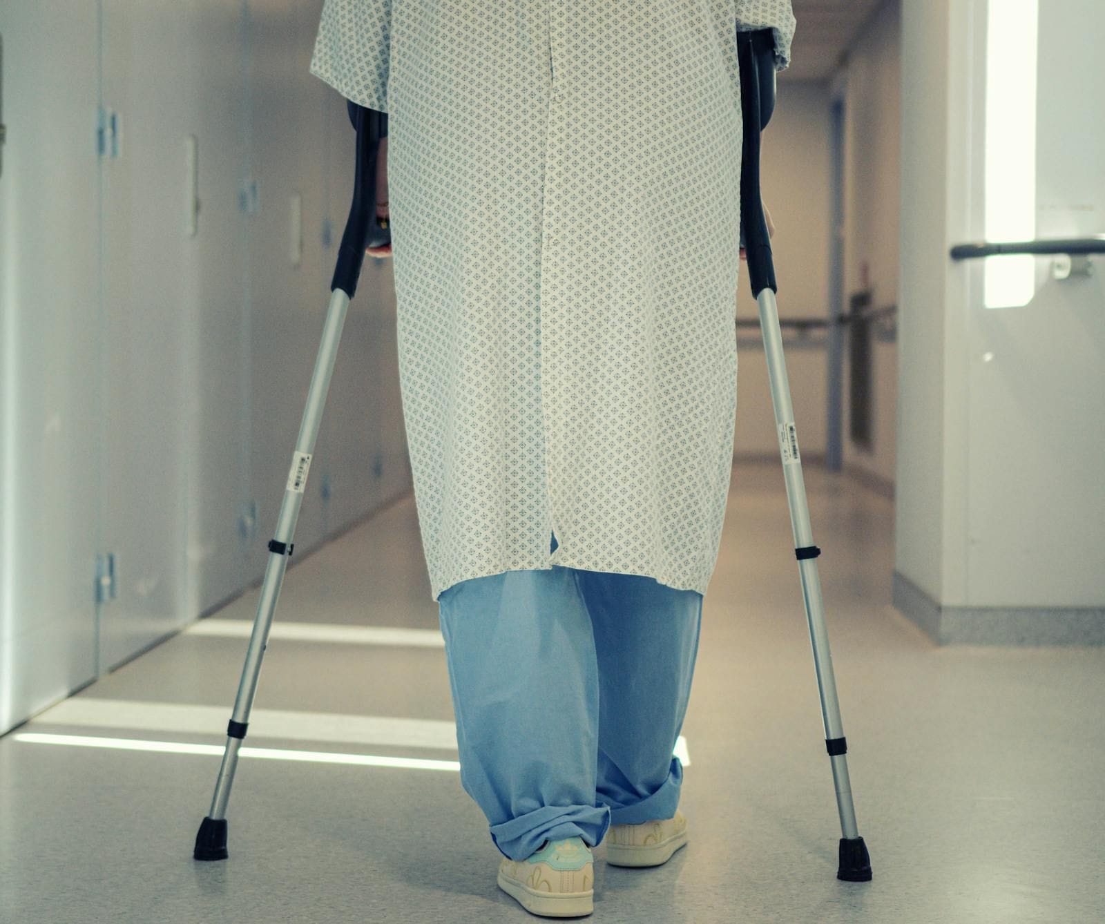 Rear view of a patient in a light blue patterned hospital gown and light blue scrub pants walking forward down a brightly lit hospital corridor using two underarm crutches, wearing cream-colored sneakers, the corridor extending into soft hospital lighting