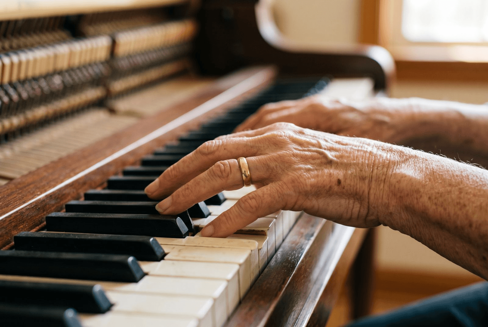 Close-up of an older person's hands resting gently on the white and black keys of a piano, wearing a simple wedding band, in soft natural light