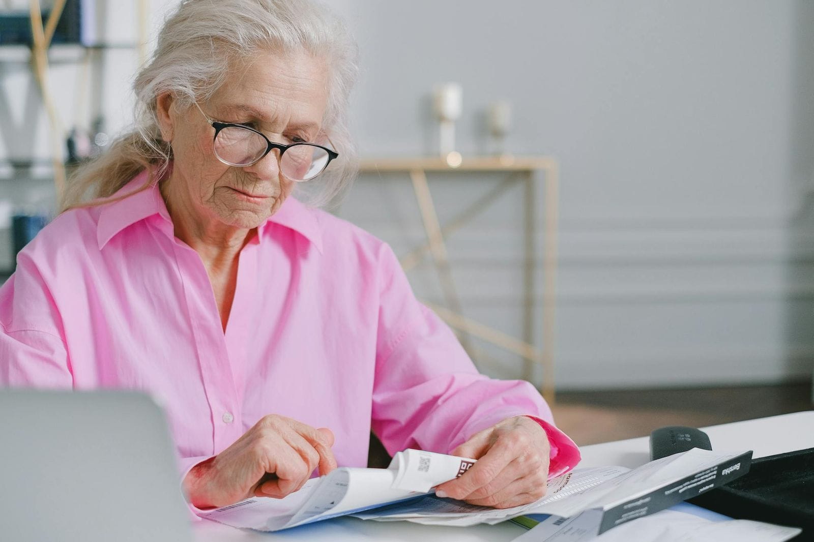 An older woman in a pink shirt reading carefully through documents at a table indoors