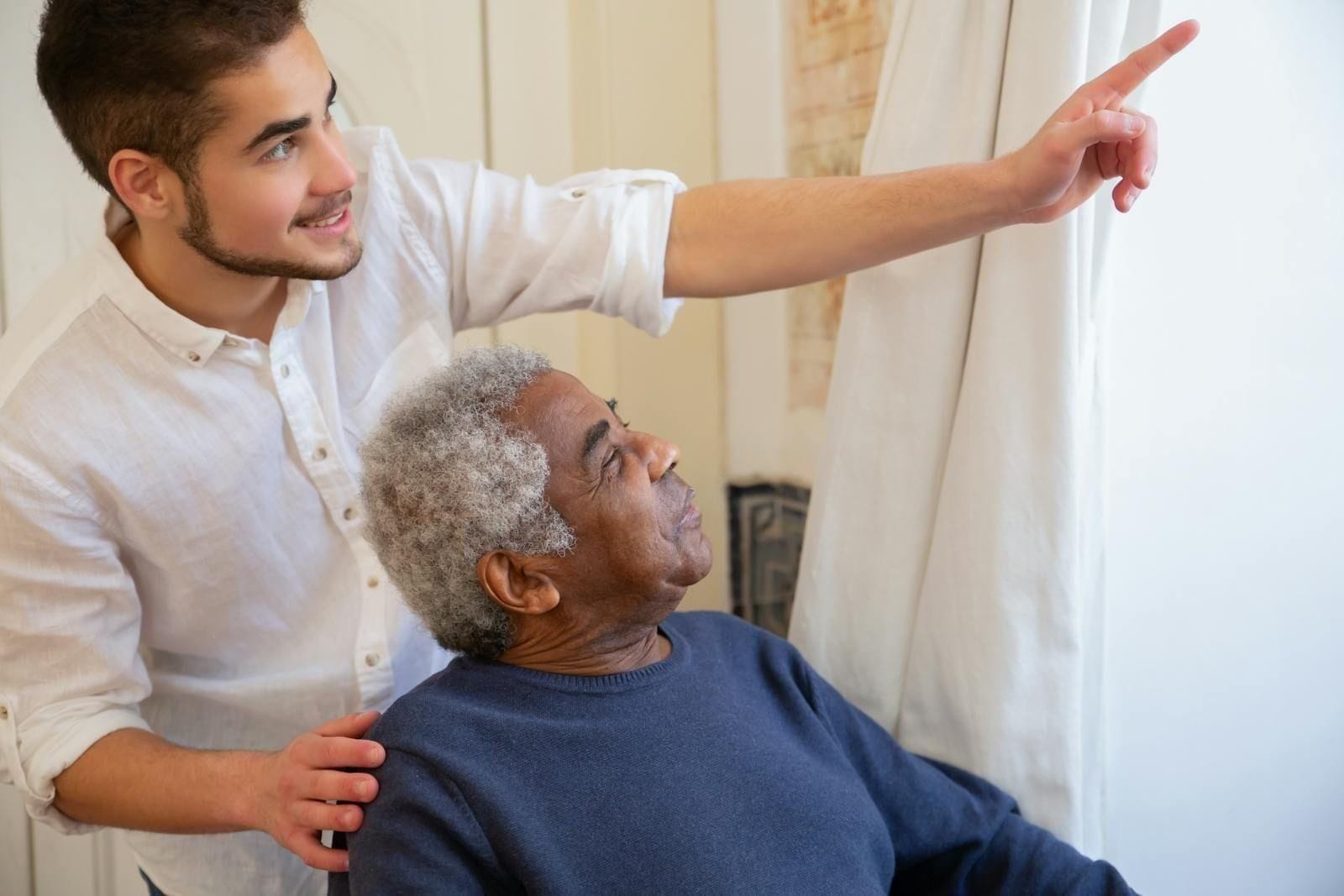 A young male caregiver in a soft white shirt stands beside an older Black man in a navy sweater seated by a window, gently resting one hand on the man's shoulder and pointing out something beyond the curtain, the two of them turned together toward the same quiet view