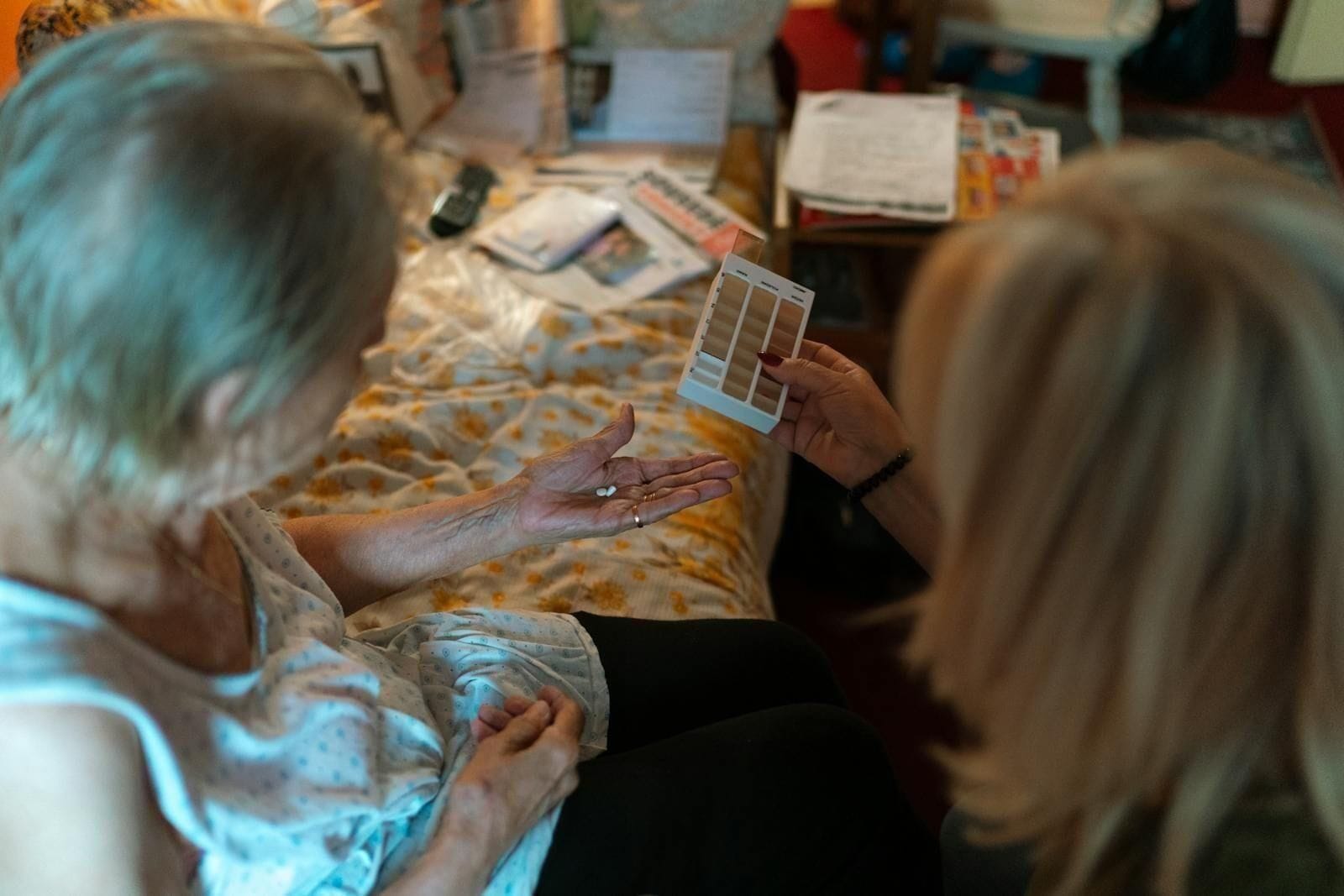 An older woman with white hair sits on the edge of a floral bedspread, palm open to receive two small white pills, as a younger woman with blond hair leans toward her holding a printed weekly pillbox, the quiet ritual of medication management between two people who clearly know each other well