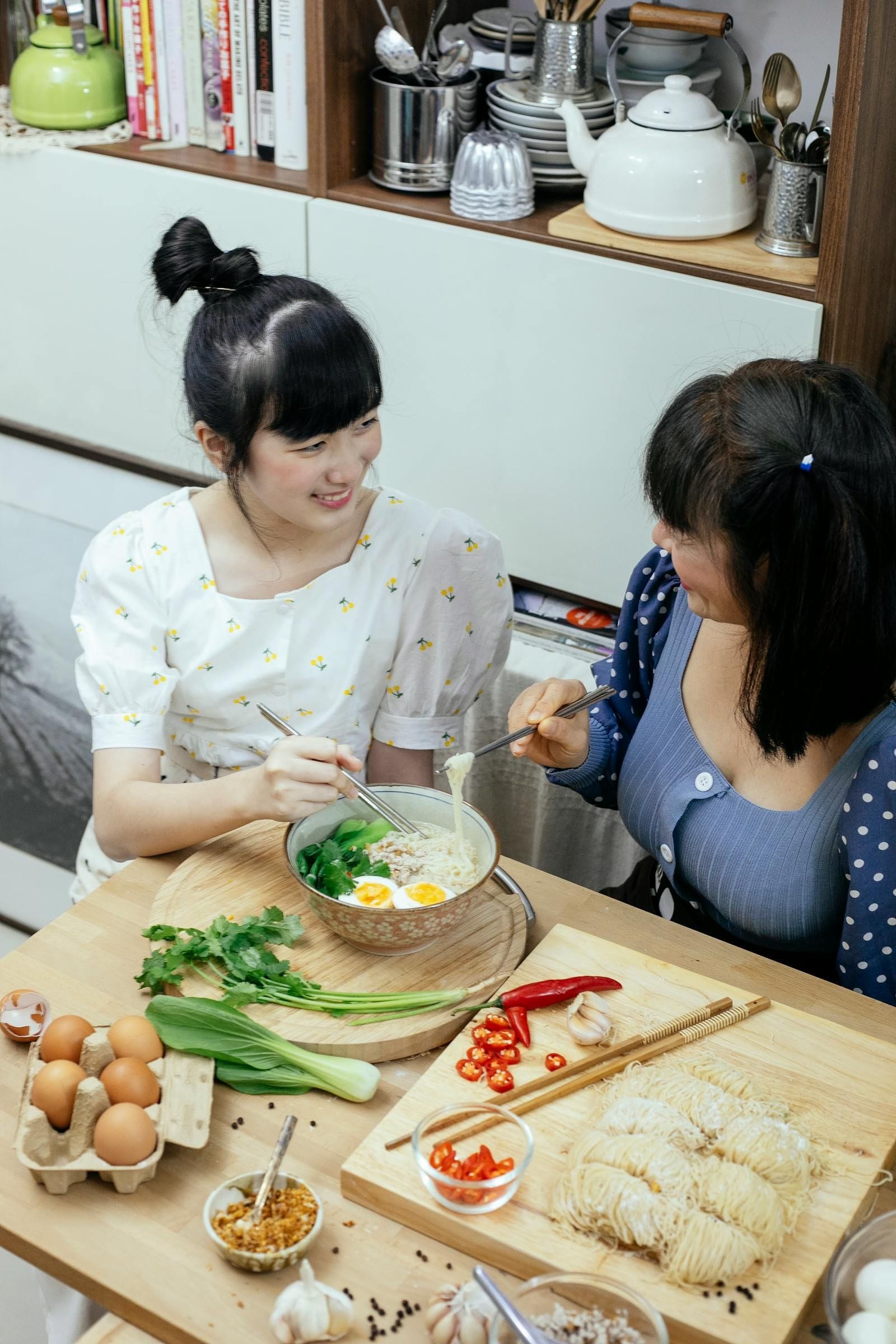 An older woman and a younger woman lean over a shared bowl of warm soup at a small table, smiling and talking with their spoons mid-air, illustrating the way company at the table often does more for an older adult's appetite than a longer ingredient list