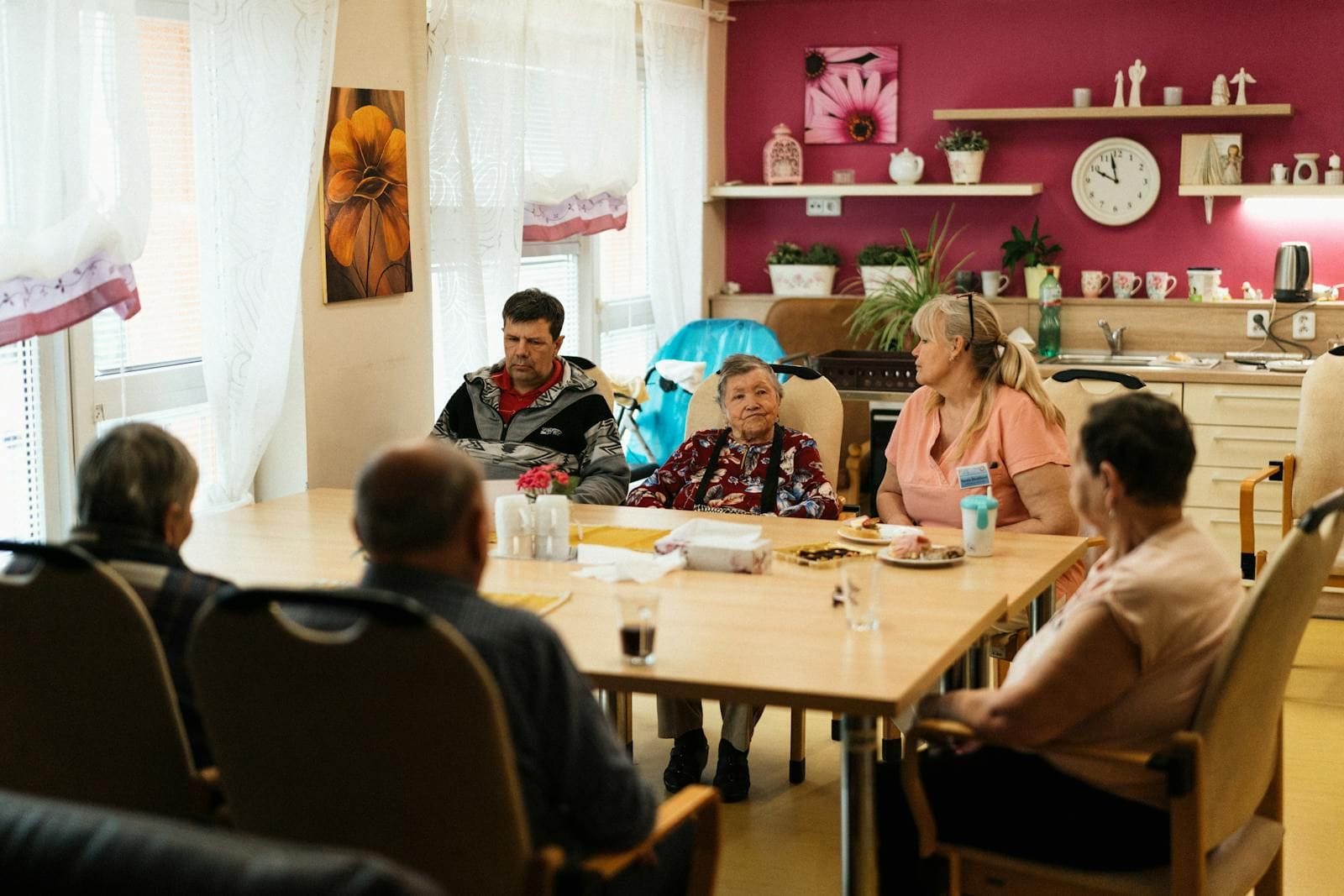 An older man in a checkered shirt sits at a small kitchen table across from a younger female caregiver, both leaning into a quiet conversation over a shared meal in a softly lit family home