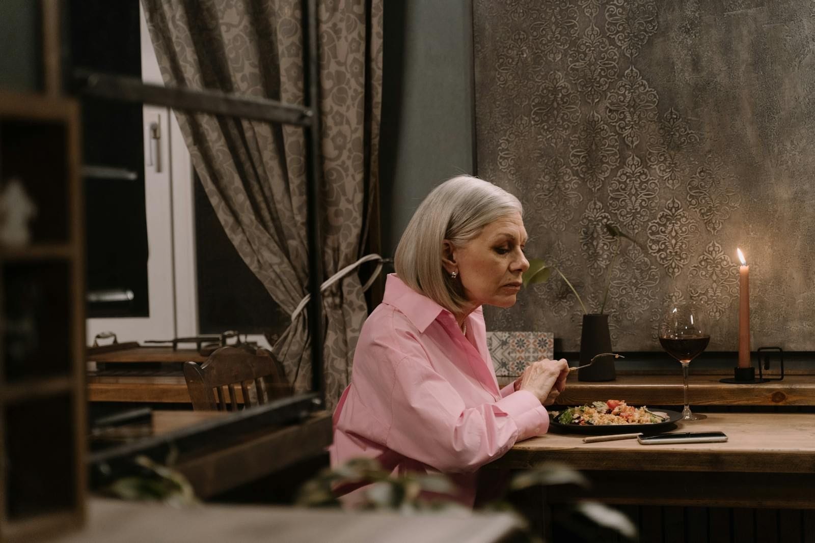 An elderly woman in a soft pink blouse sits at a wooden table beside a lit candle, holding a fork above a half-finished plate of food, gazing thoughtfully off to one side as warm window light falls across the kitchen behind her