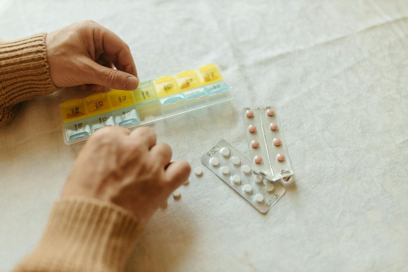A person in a warm knit sweater sorting tablets from blister packs into a seven-day pill organizer at a table