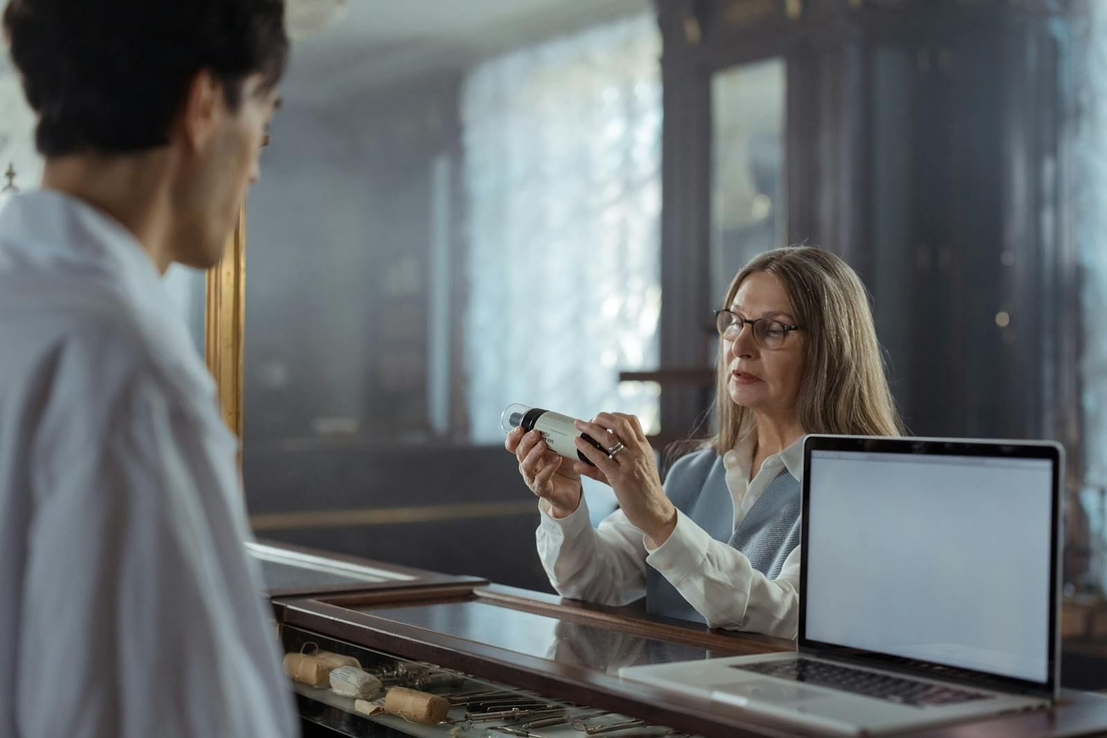 A silver-haired woman in glasses reading the label on a medicine bottle at a pharmacy counter