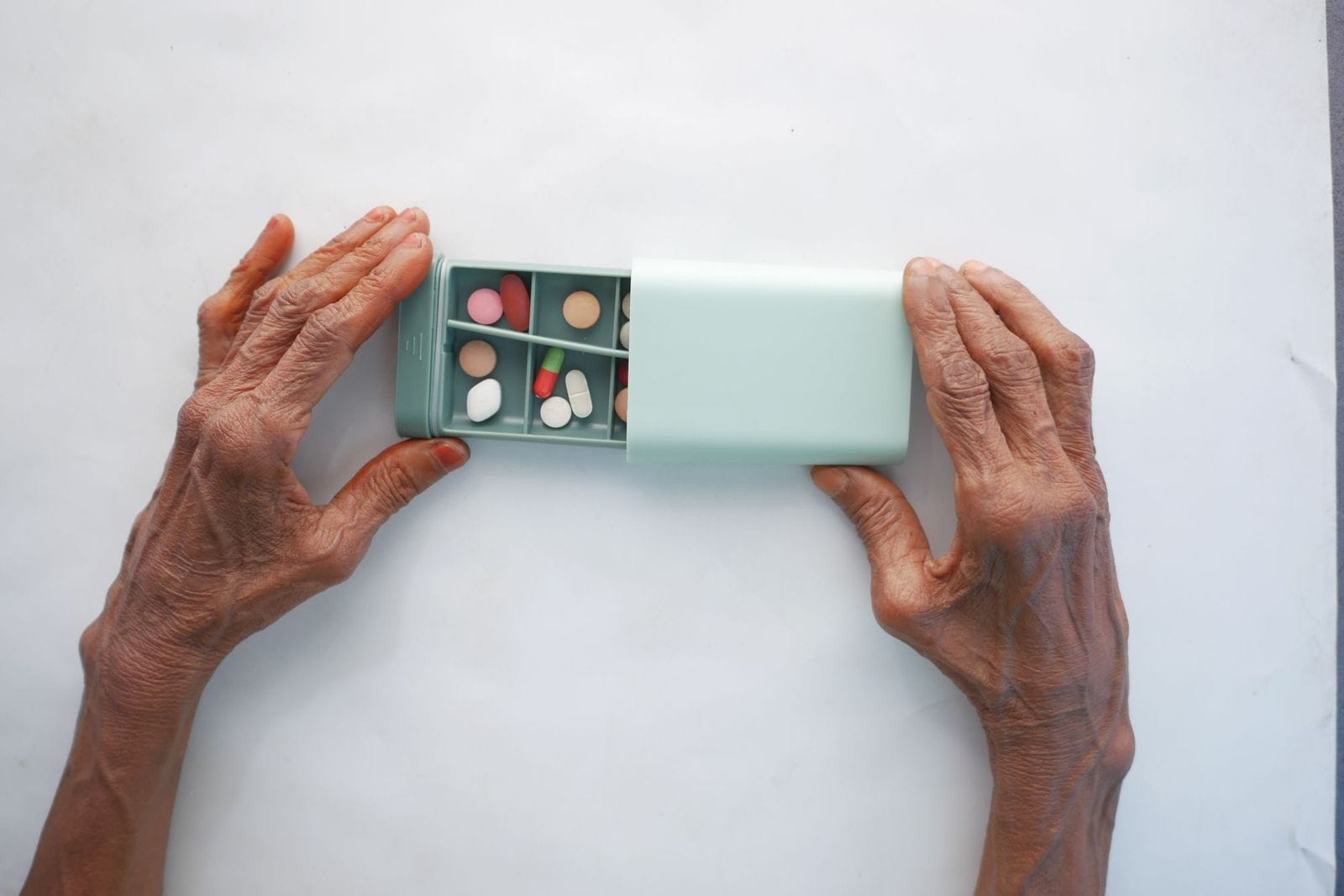 An older adult's hands opening a pale green weekly pill organizer filled with assorted tablets and capsules on a marble surface