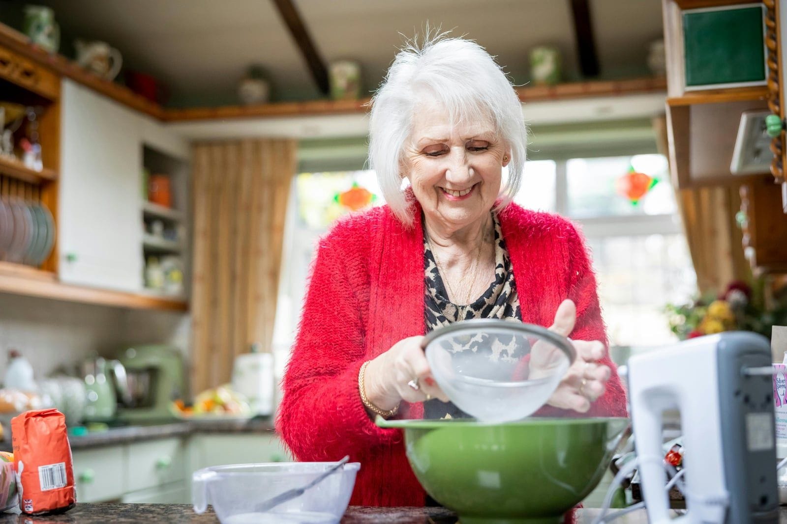 A white-haired senior woman in a red cardigan stands smiling in her own sunlit, cluttered home kitchen, sifting flour into a green mixing bowl, surrounded by familiar shelves, plants, and the cheerful disorder of a house lived in for decades