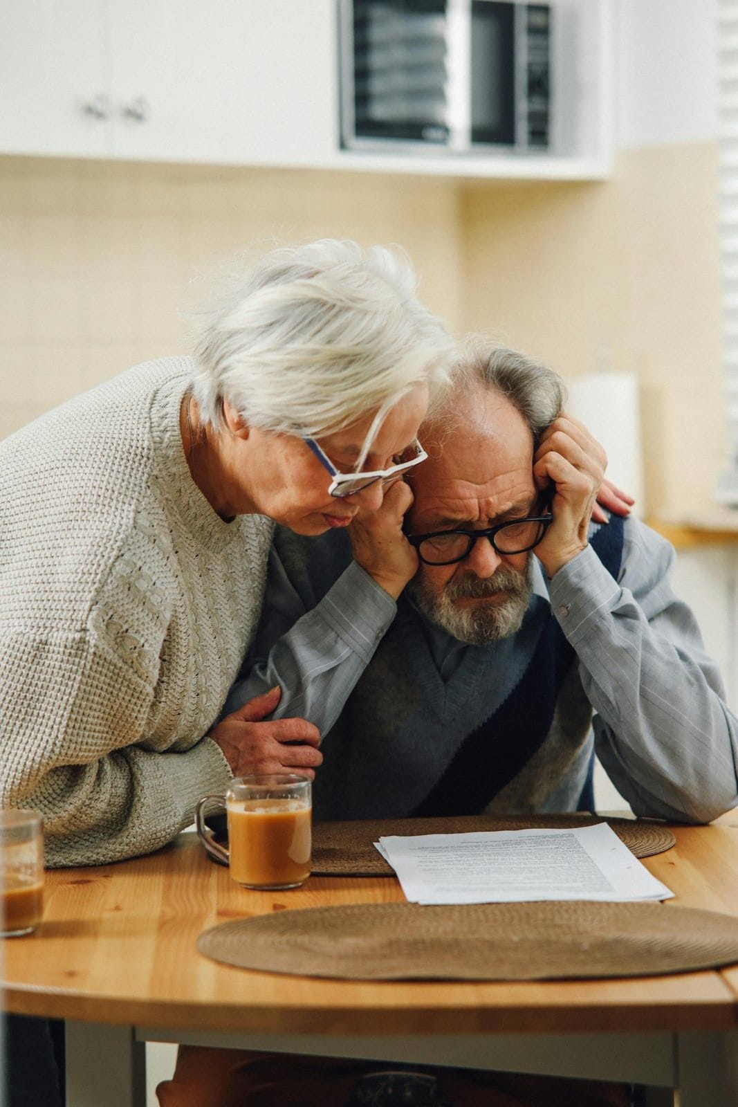 A senior couple at a kitchen table reviewing a sheet of paperwork together, the wife in a cream sweater leaning in with one hand on her husband’s shoulder while he sits in a vest with his head in his hands, a glass of coffee untouched beside the documents